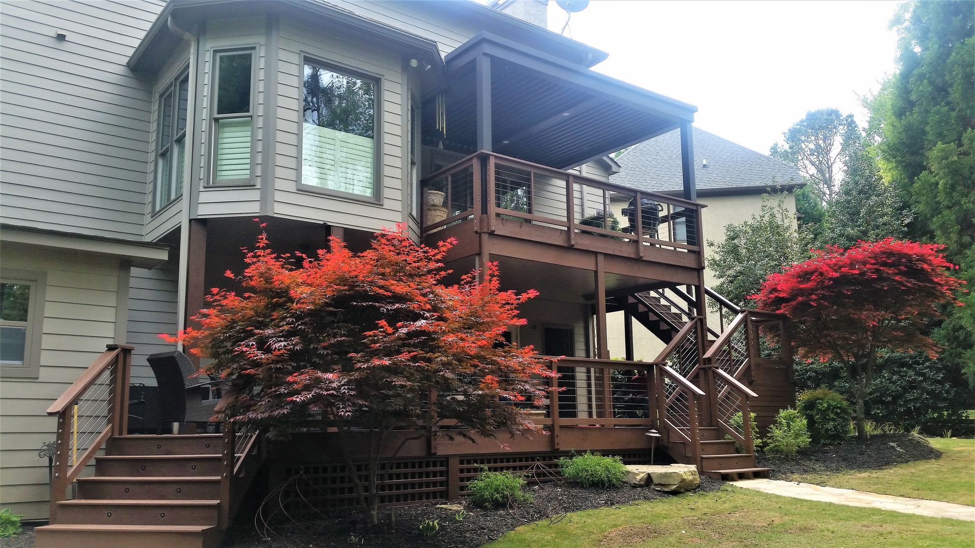 Two-story wooden deck attached to a house with bay windows; red shrubs in the foreground.