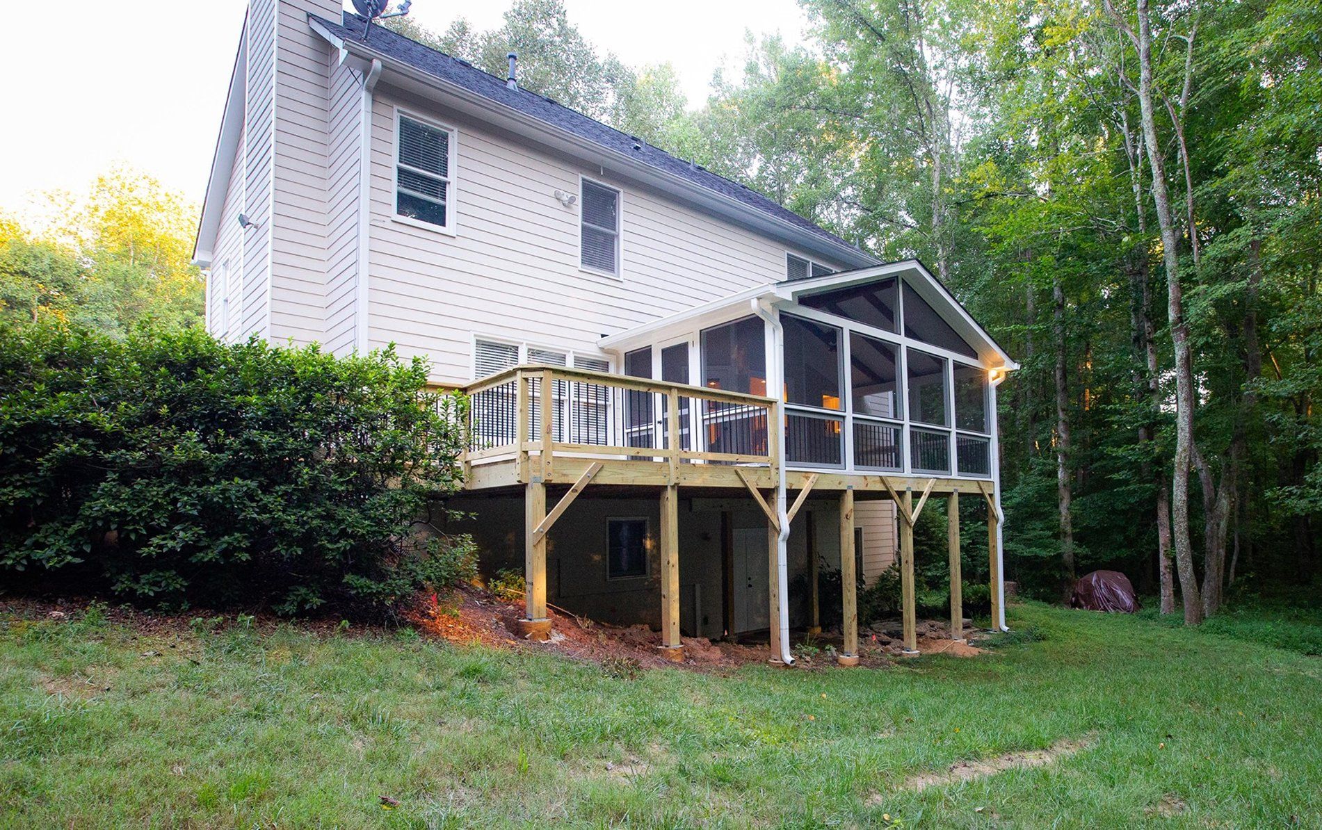 Two-story house with wooden deck and screened porch, surrounded by green grass and trees.