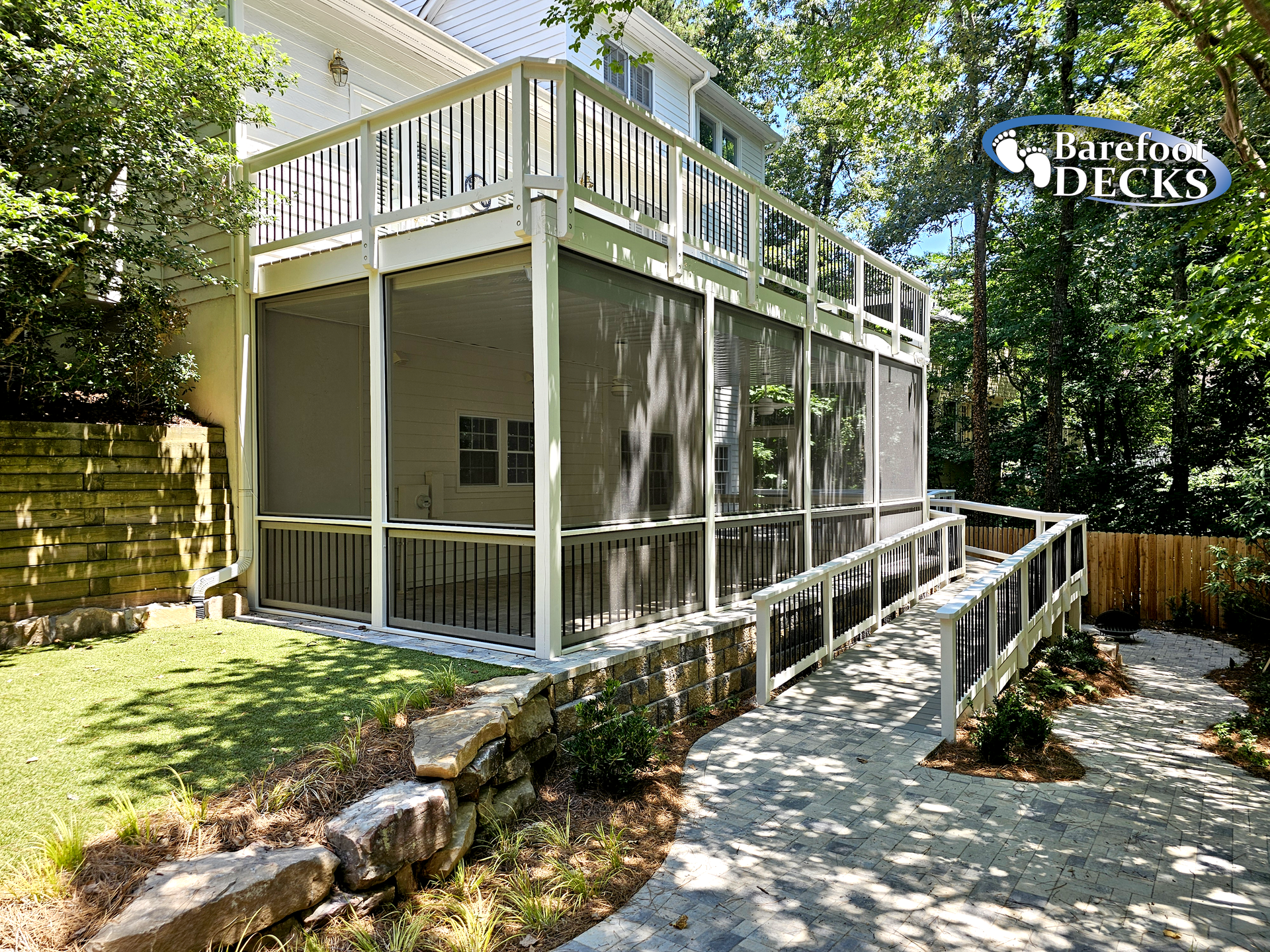 A multi-level white deck with a screened porch and ramp, built next to a house, in a wooded backyard.