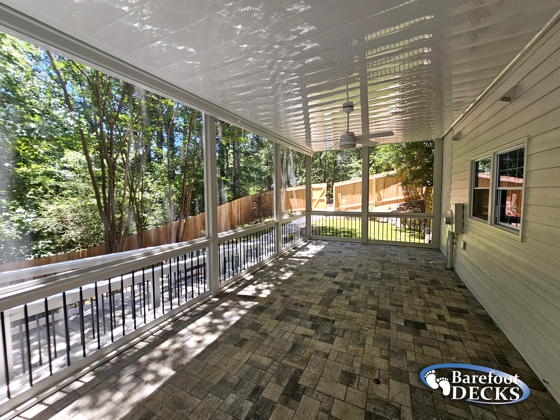 Screened-in porch with brick-patterned floor, overlooking a wooded backyard. White walls and railing.