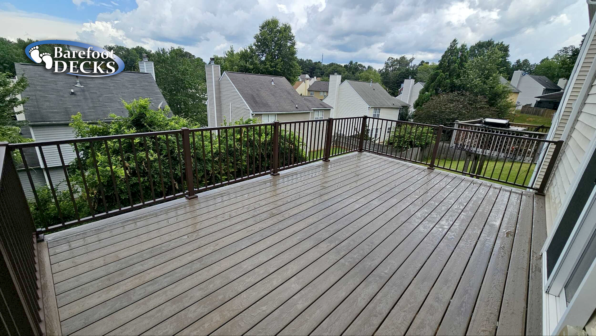 Wooden deck with black railing overlooking a residential area with green trees and houses.