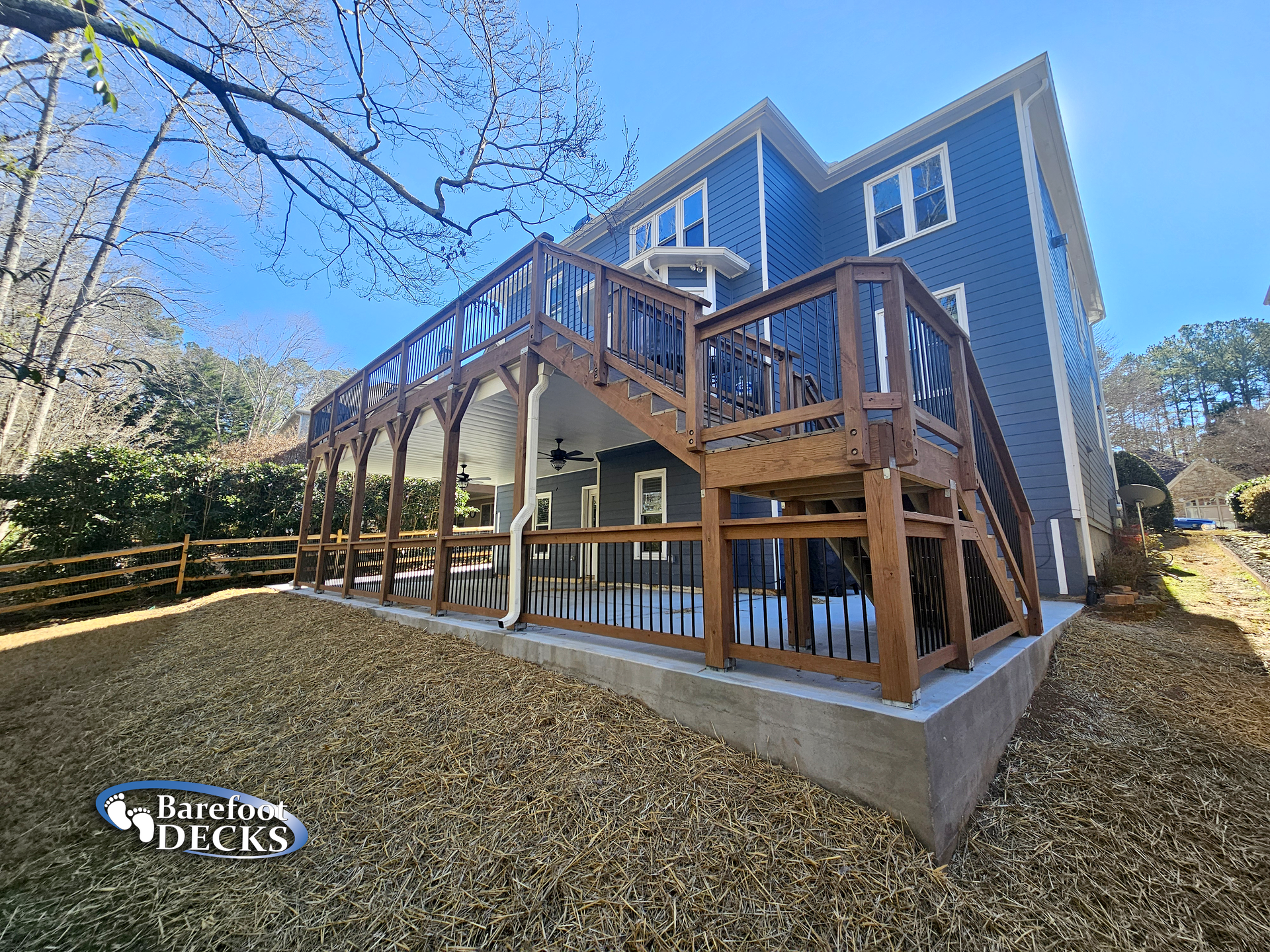Wooden deck with stairs attached to a blue house, in a yard with wood chips and a fence.