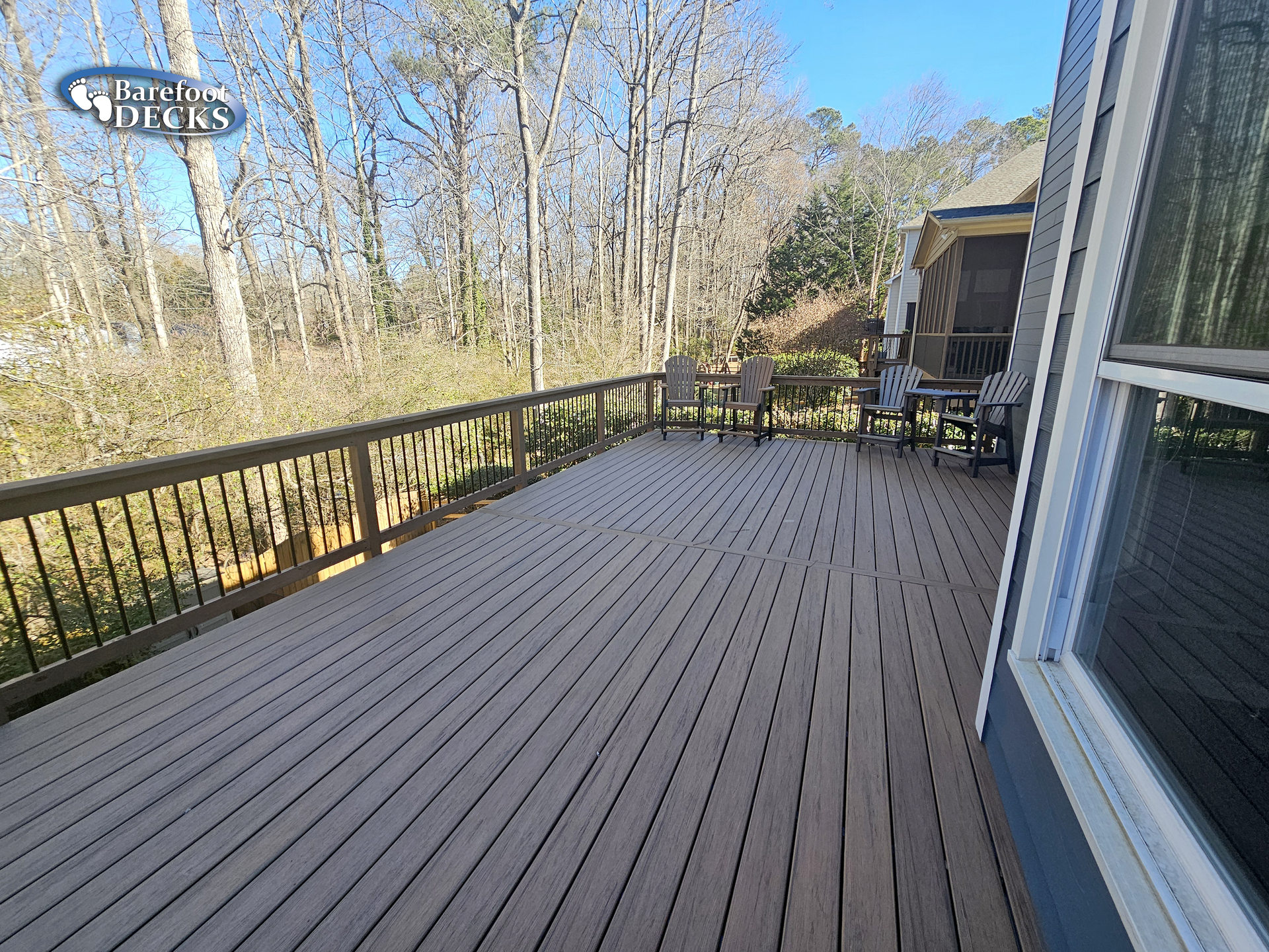 Wooden deck with railing, chairs, and trees in the background. Bright, sunny day.