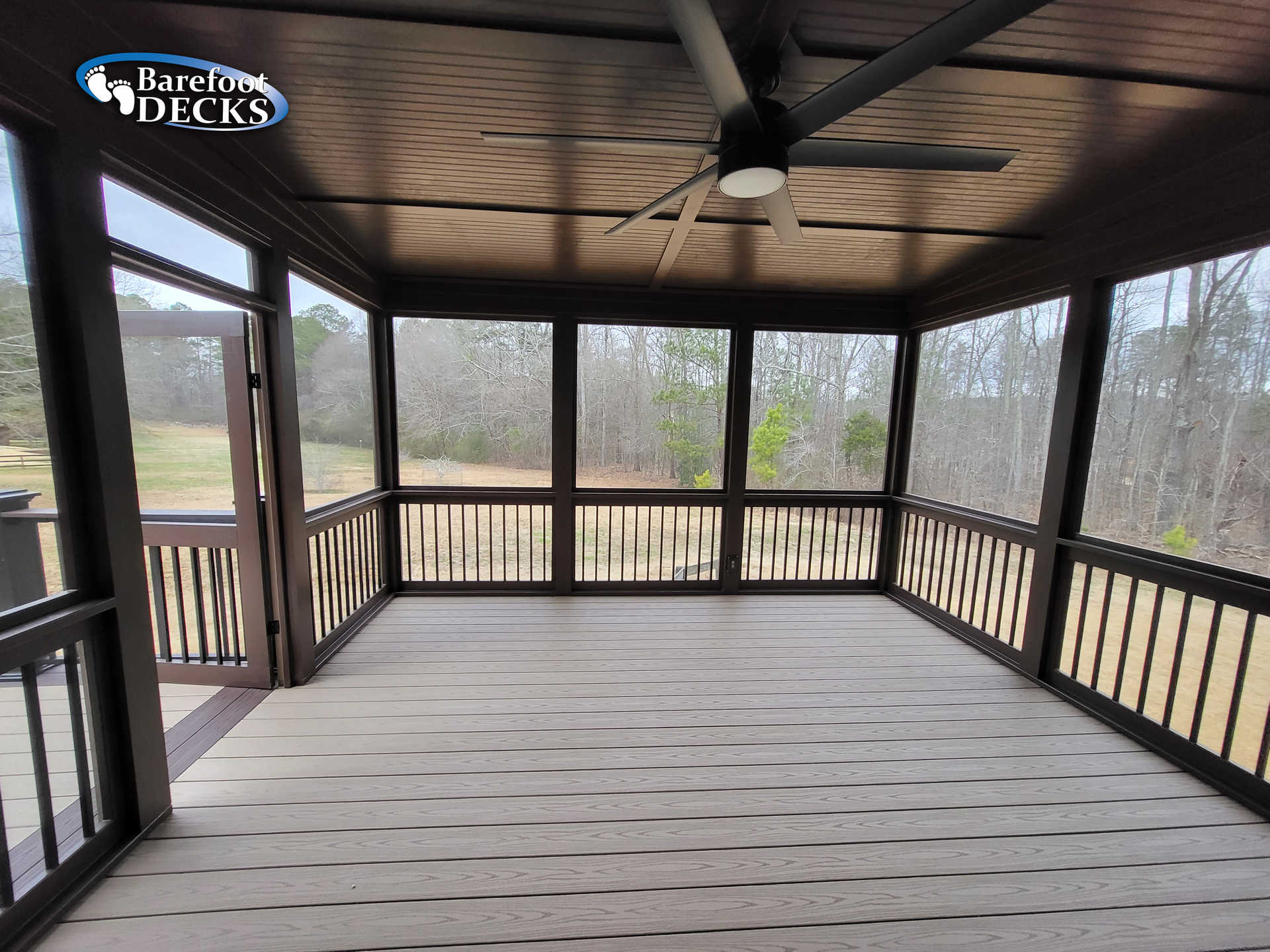 Brown screened-in porch with light gray deck, dark ceiling, and a ceiling fan, overlooking a wooded area.
