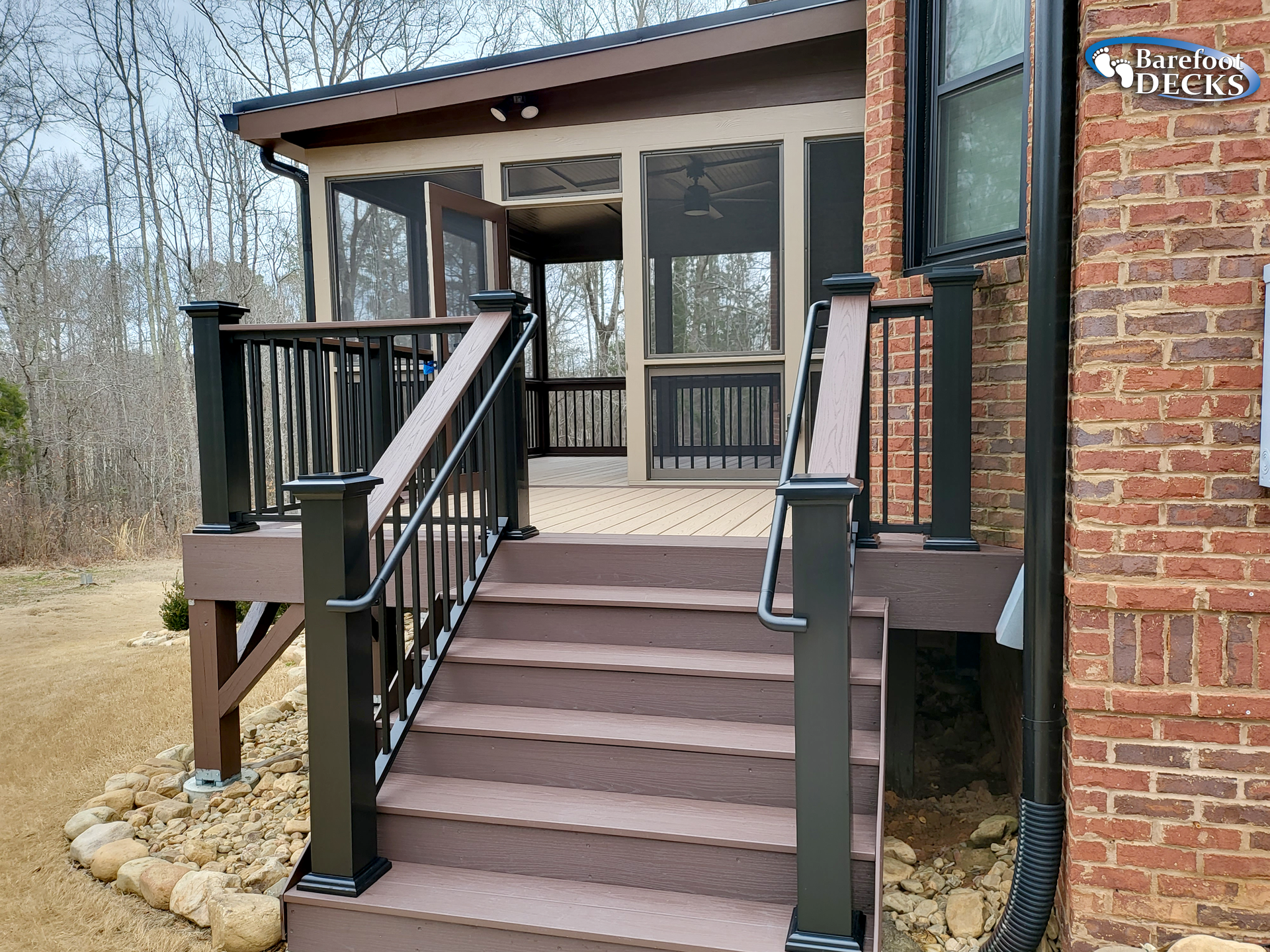 Brown deck with steps leading to a screened porch, black railings, attached to a brick house.