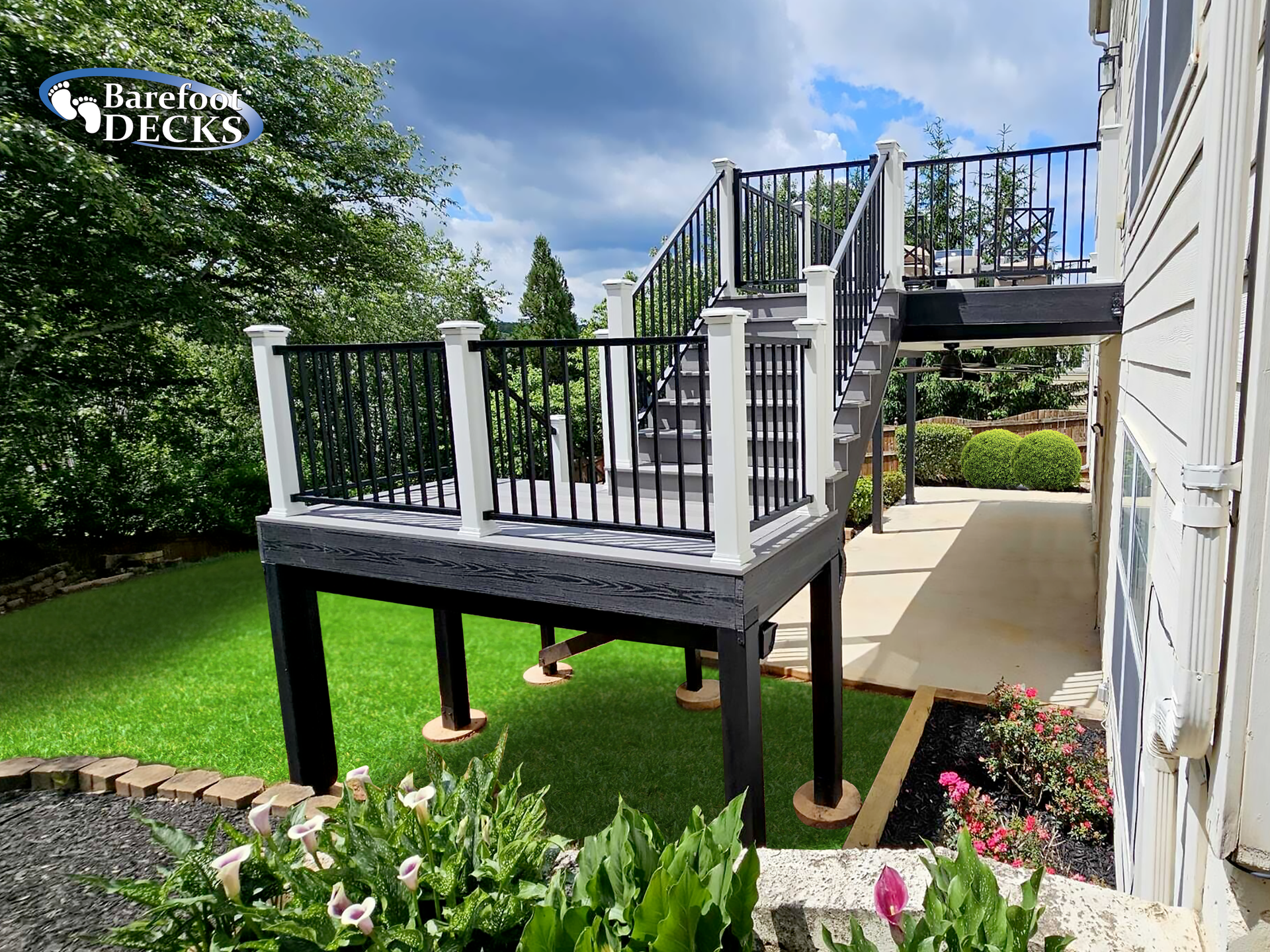 Deck with black railing, white posts, and stairs, next to a house with a green lawn.