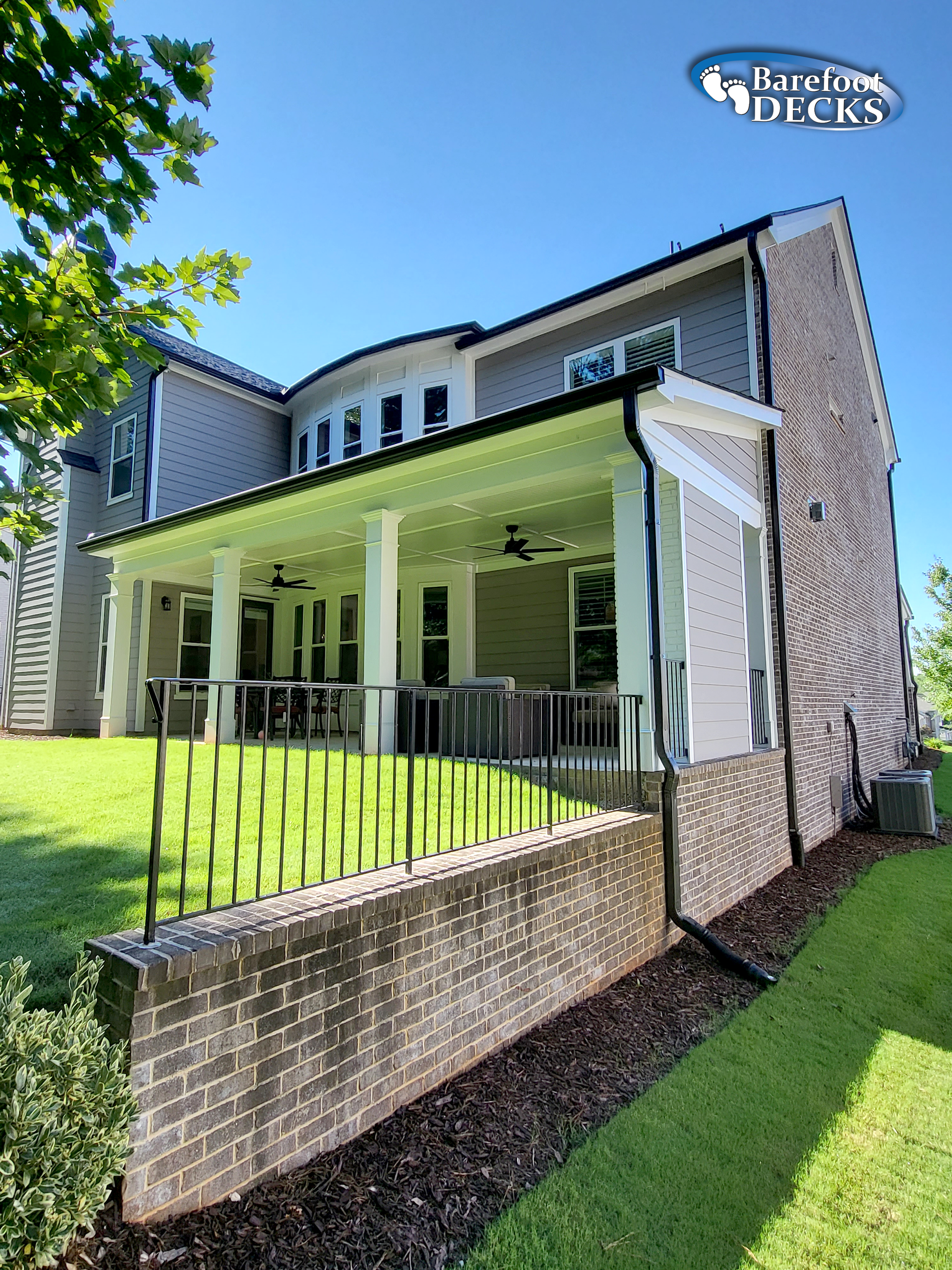 Back of a house with a porch, black railing, brick wall, and green lawn on a sunny day.