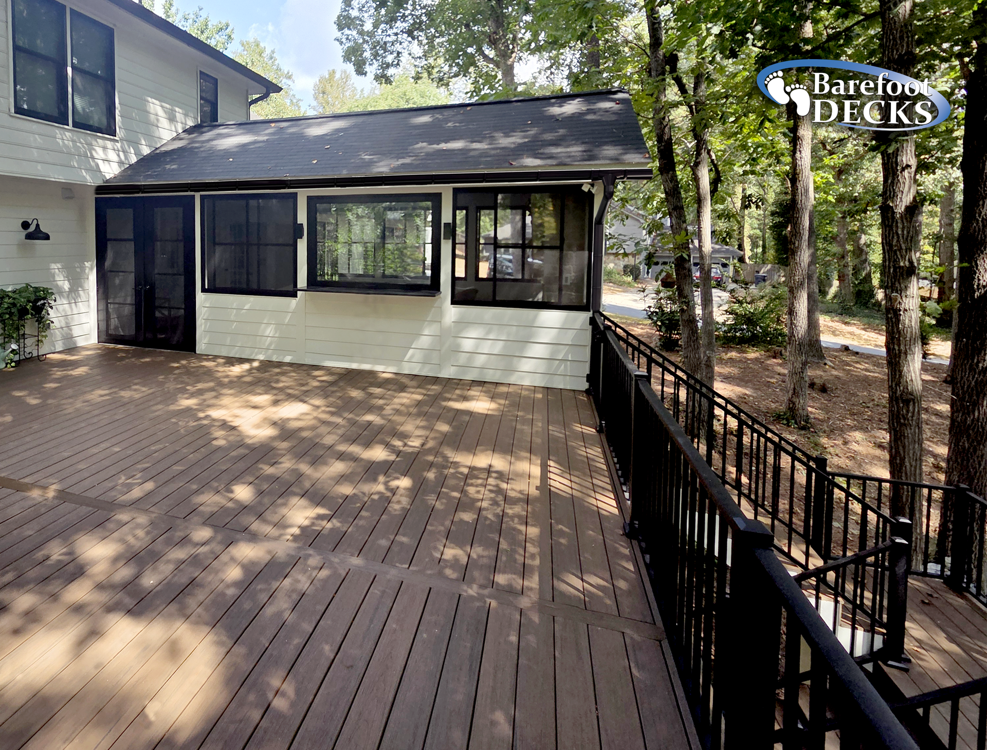 Composite deck with black railing and screened porch connected to a white house, surrounded by trees.