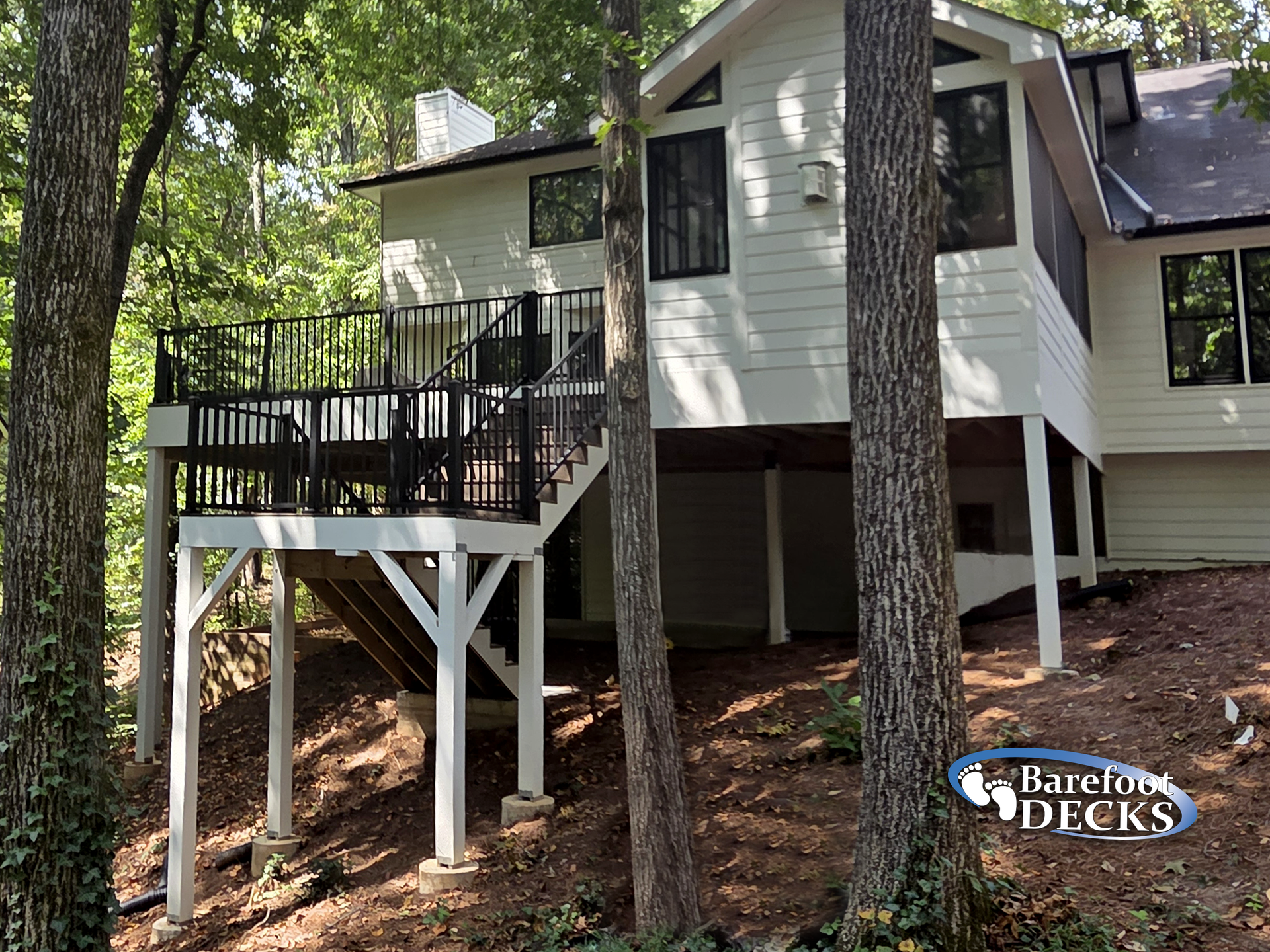 Deck of a white house with black railings, supported by white posts, in a wooded area.
