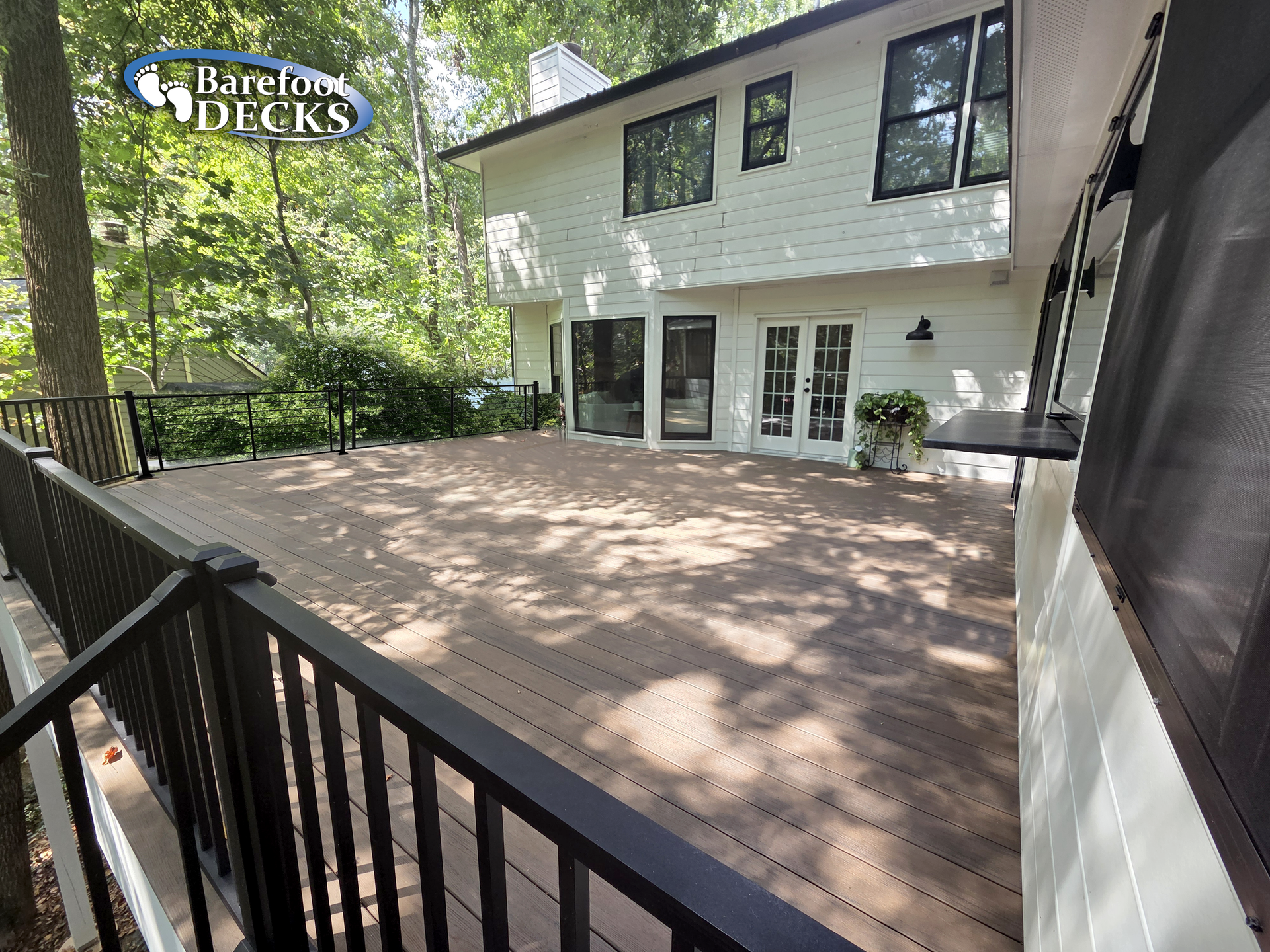 Large brown composite deck with black railing next to a white house with large windows and a wooded backdrop.