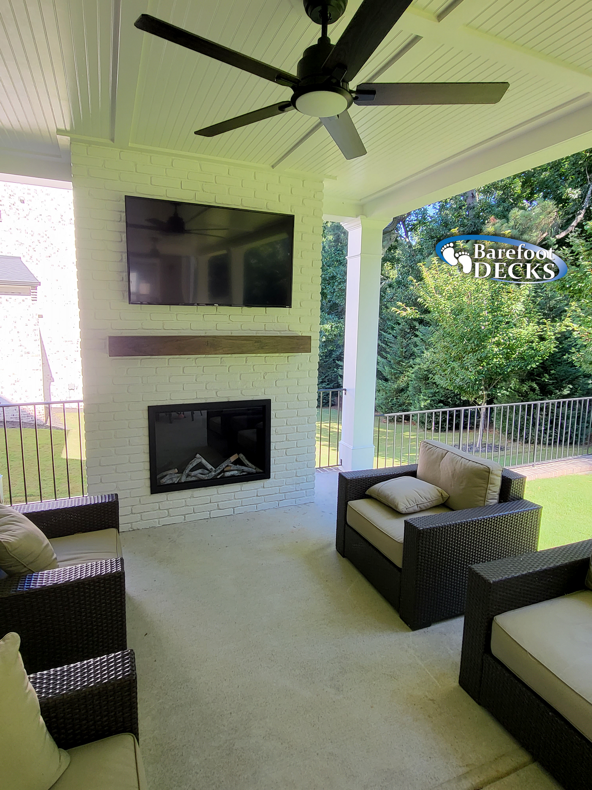 Covered patio with fireplace, TV, seating; white brick, ceiling fan, view of backyard.