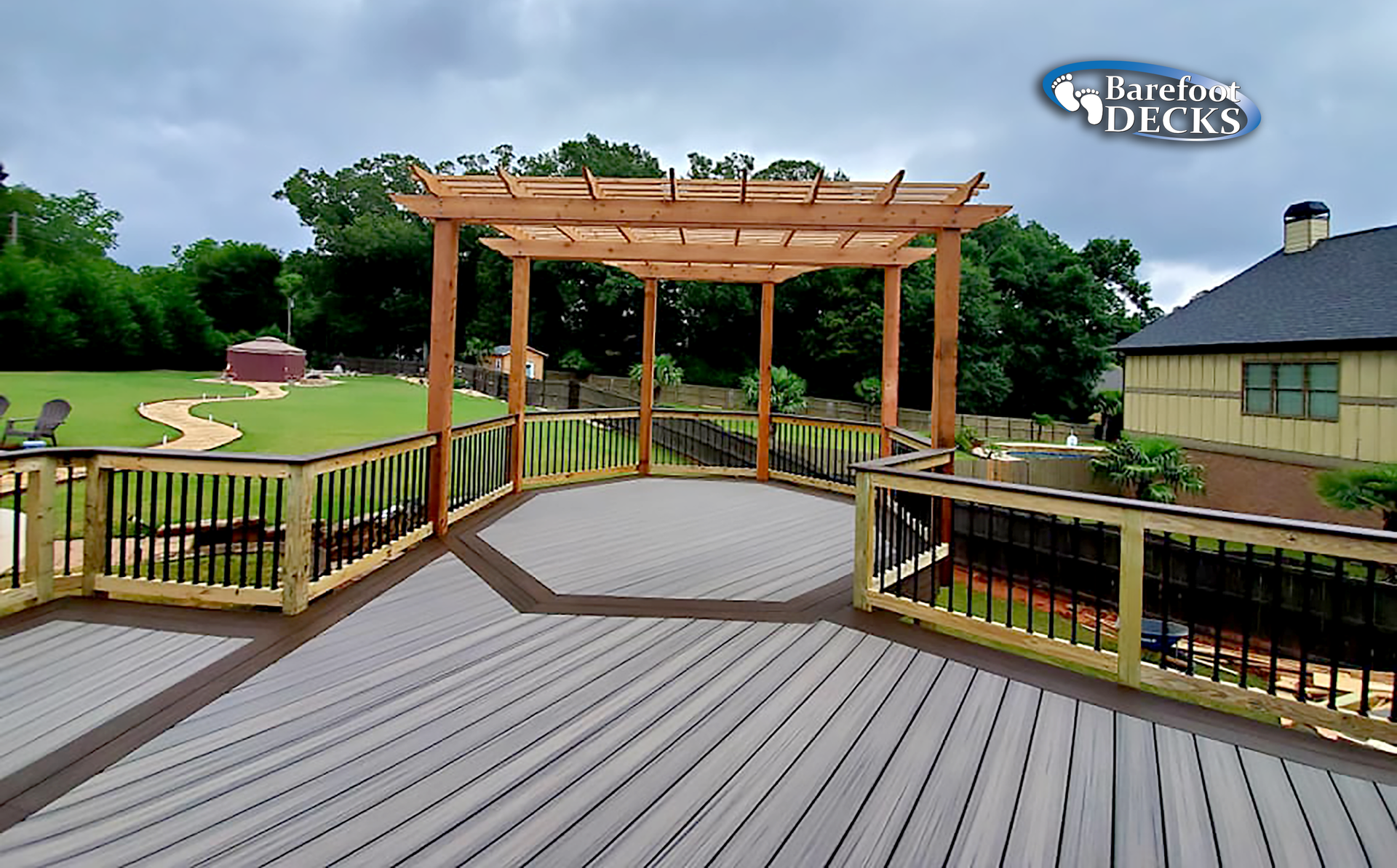 A wooden deck with a pergola, brown railings, and a view of a yard and house.
