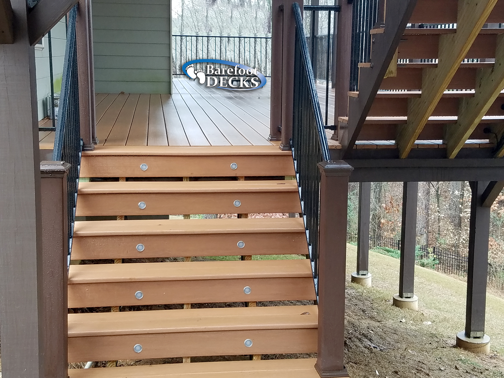Wooden deck stairs with built-in lights, brown railings, and a view of a wooded area.