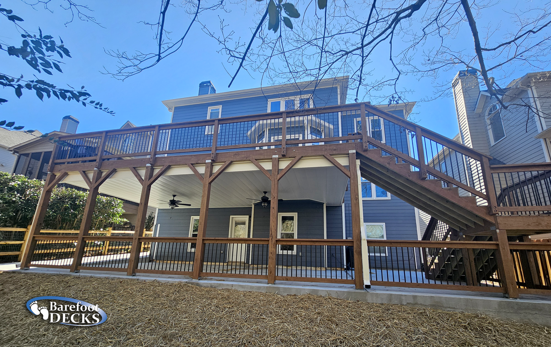 Two-story house with brown deck. Stairs lead to the upper level. Fencing surrounds the lower level. Blue sky in the background.