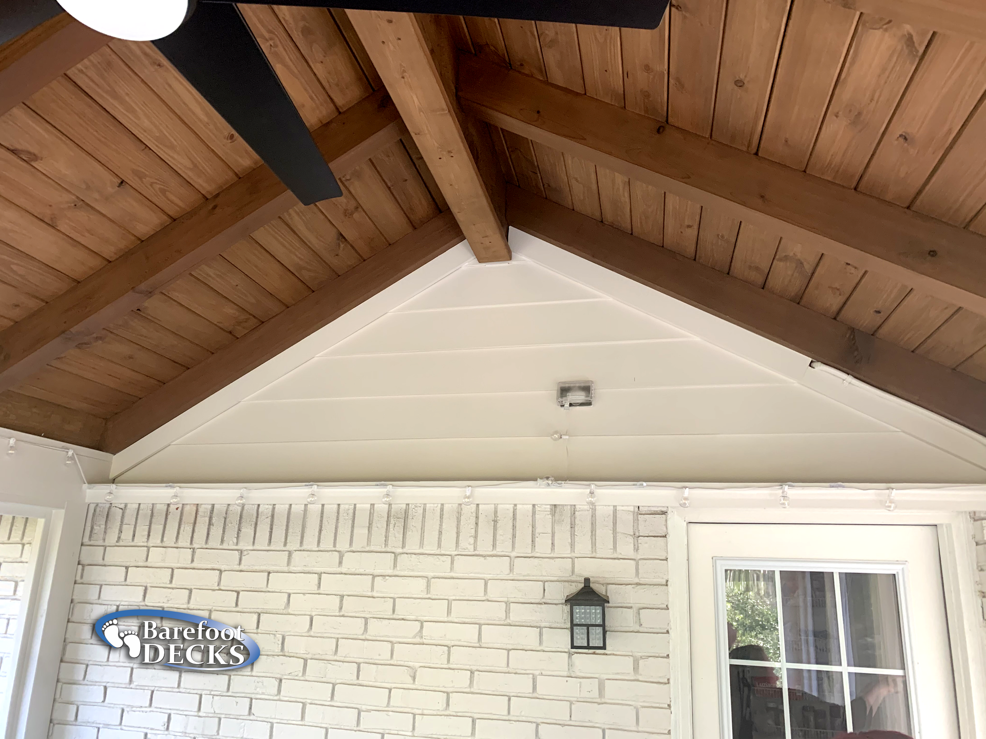 White-painted brick wall and gable with a wooden ceiling and a black ceiling fan, on a porch.