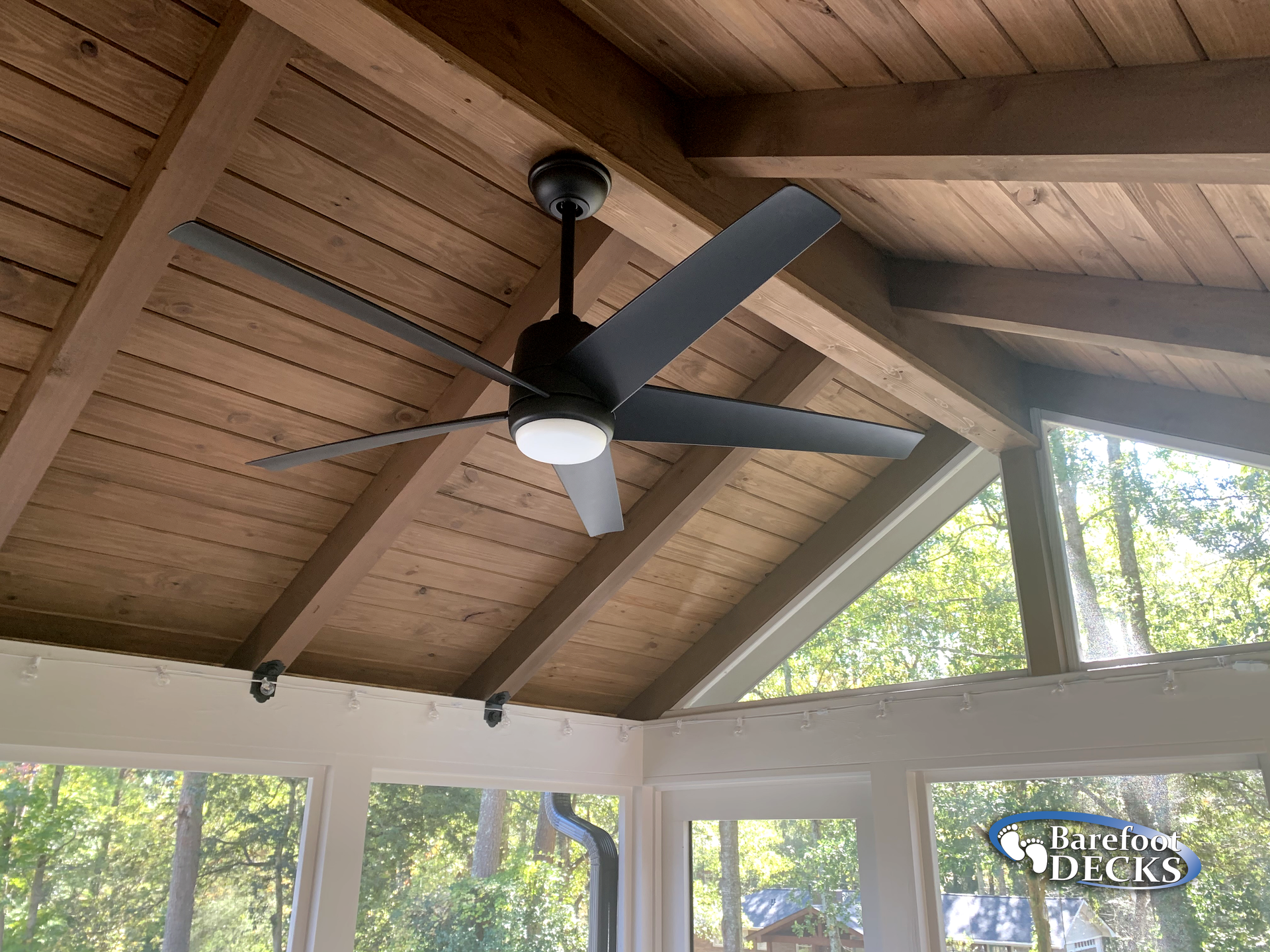 Black ceiling fan with light installed on a wooden paneled porch ceiling, with windows and trees visible outside.
