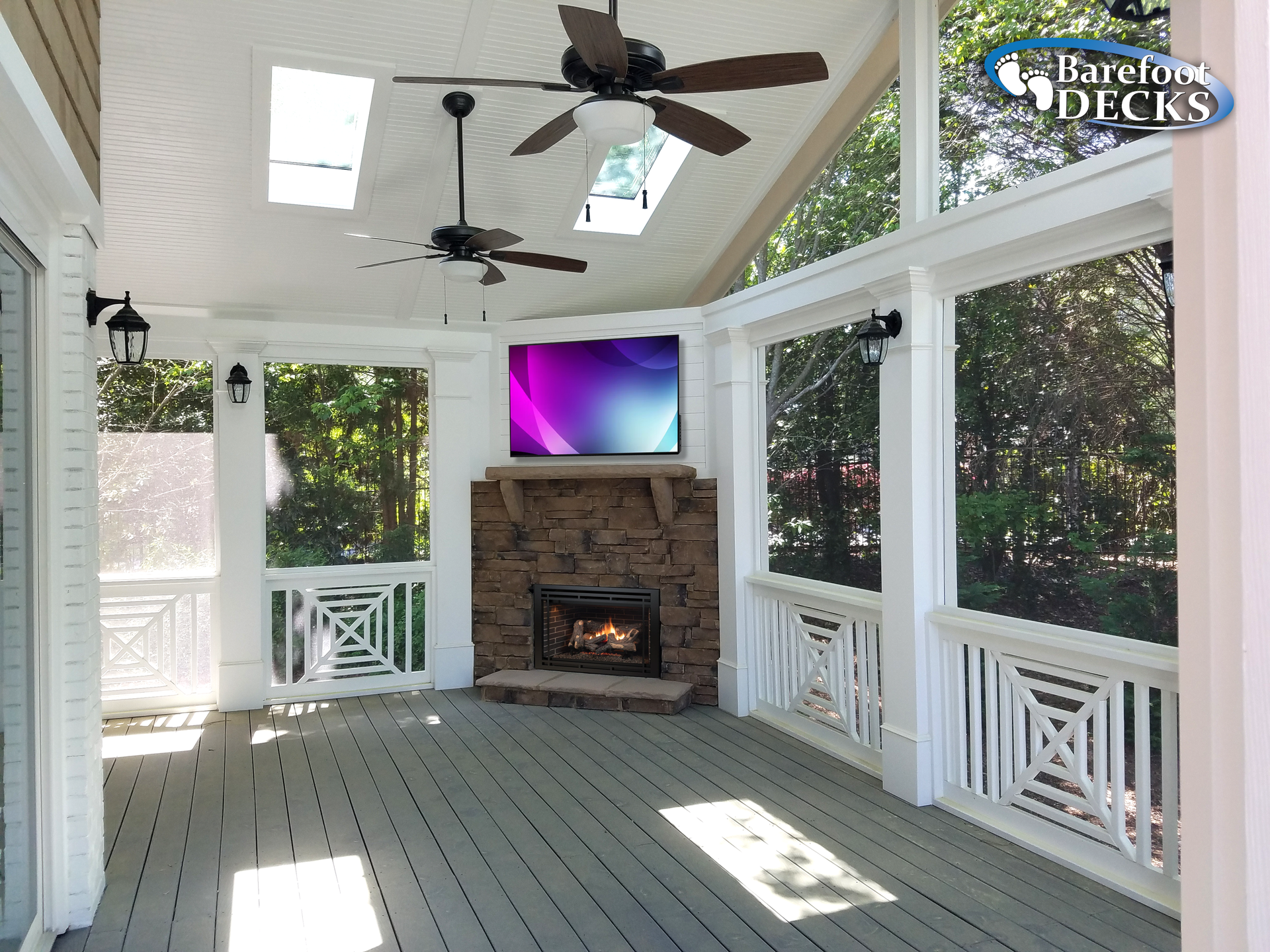 Enclosed porch with fireplace, TV, ceiling fans, and skylights.  White trim, gray floor, and views of trees.