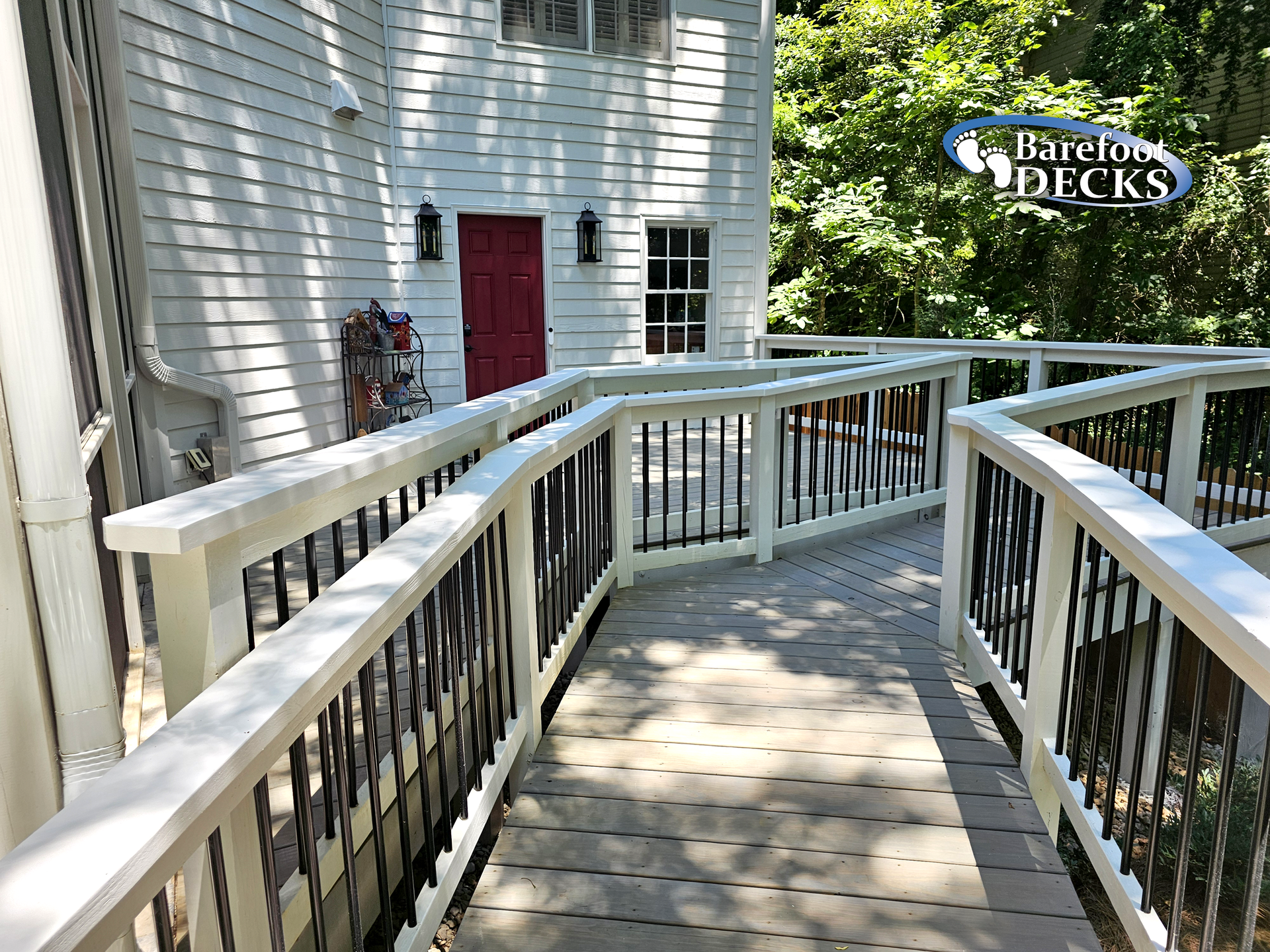 Ramp with white railing and black spindles leading to a red door on a white house.