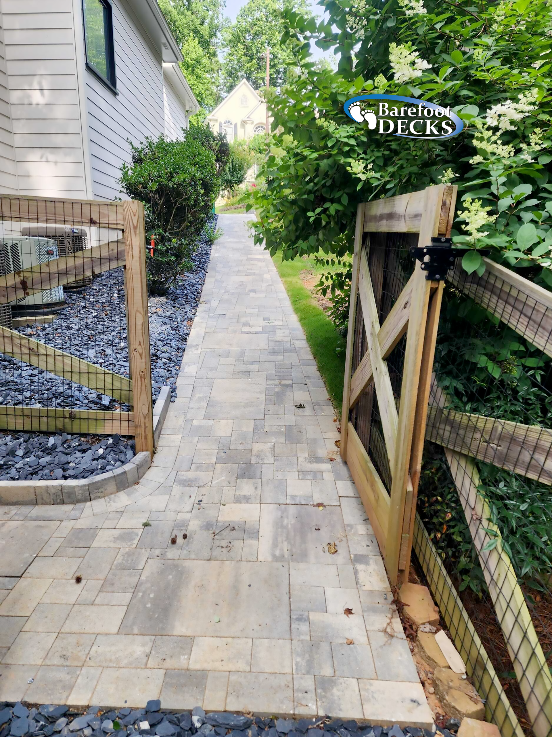 Paver pathway between fences and bushes leads to a house; partially visible is a wooden gate.