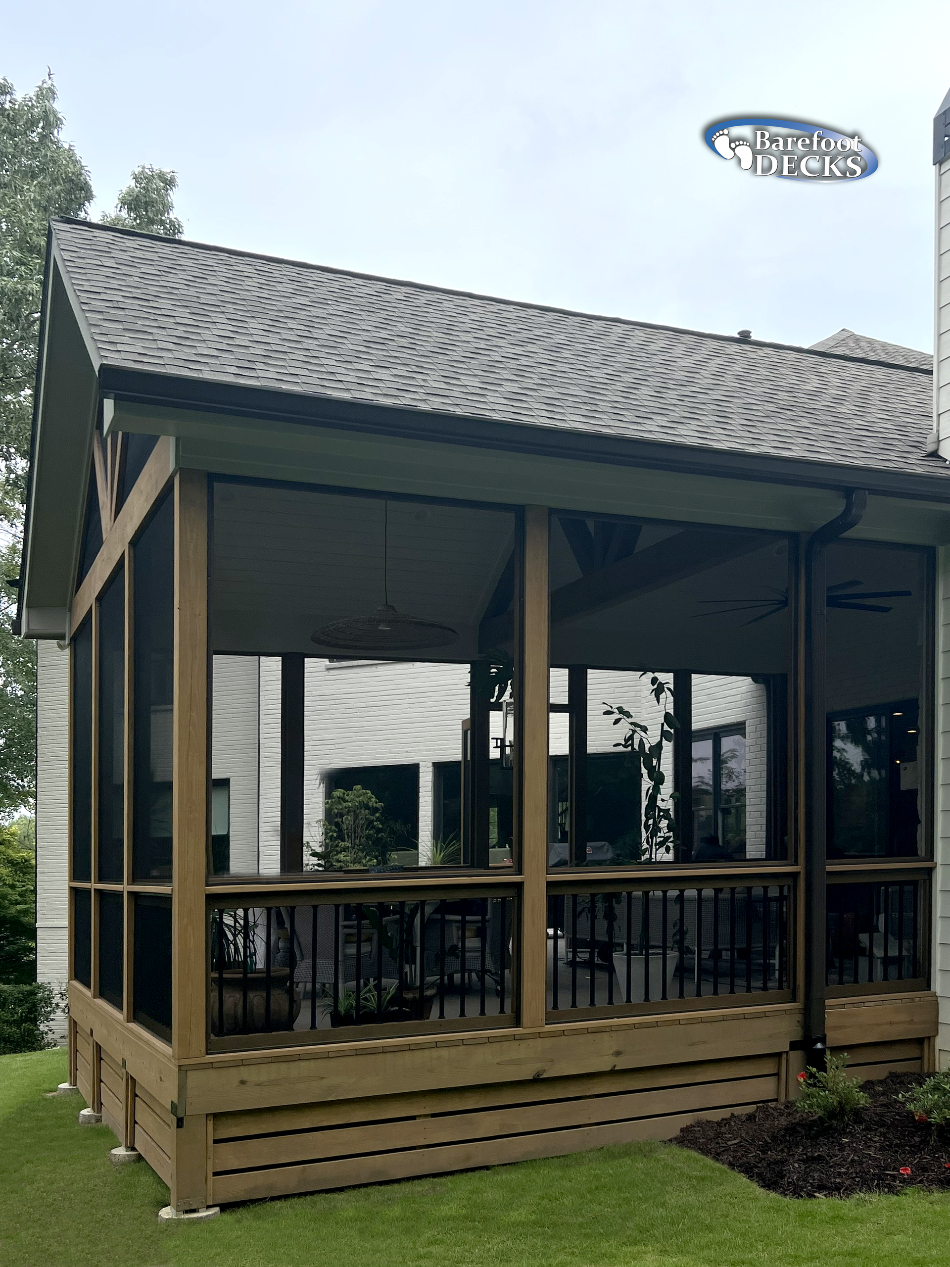 Screened-in porch attached to a house with a shingled roof, brown wooden deck, and black railing.