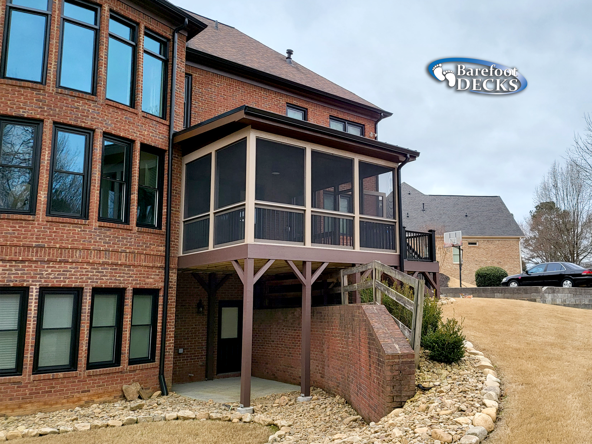 A screened-in porch with brown frame attached to a brick home. Includes a deck, stairs and door.