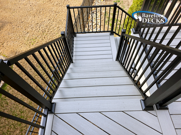 Narrow gray deck stairs with black railings leading down to a tan yard.