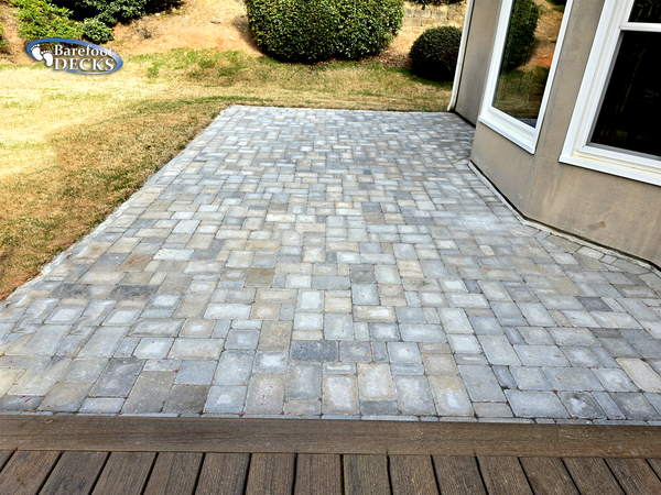 Gray stone patio beside a house, bordered by dry grass and shrubs.