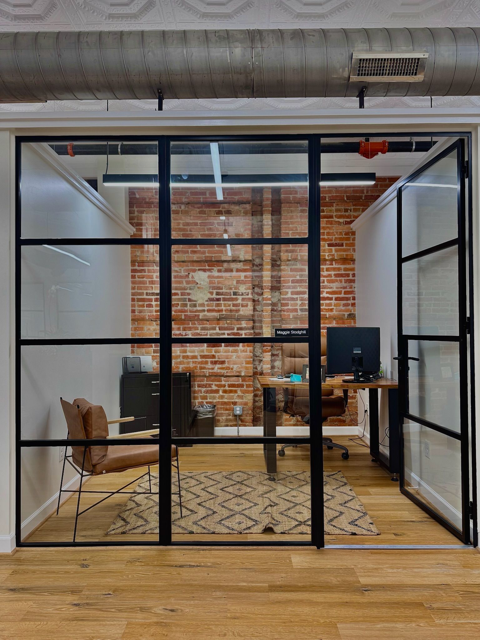 Office with glass walls and black frames, brick interior, wooden floor, and a desk.