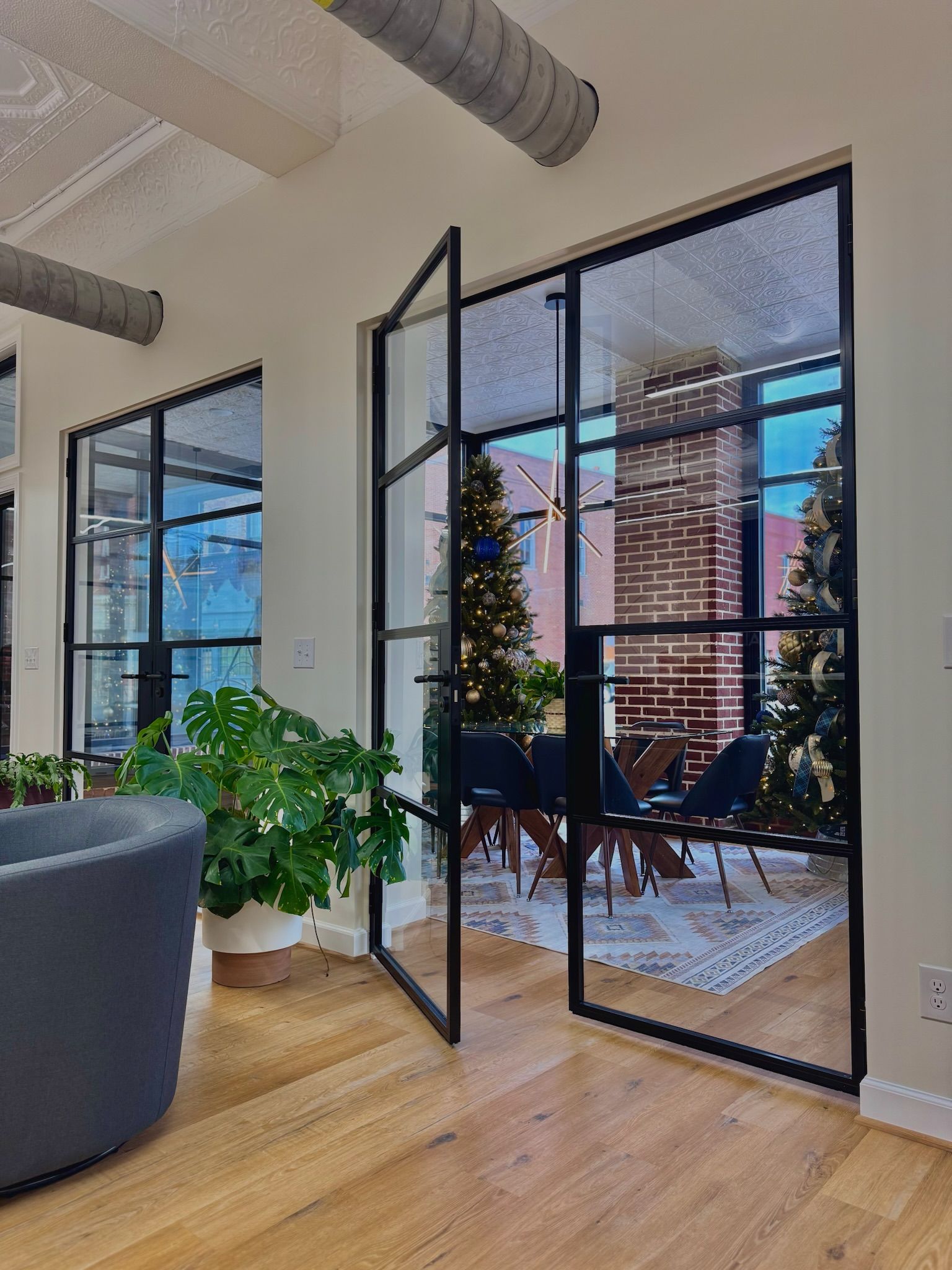 Black framed glass doors open to a dining area. A Christmas tree is visible. Wooden floors and plants are in view.