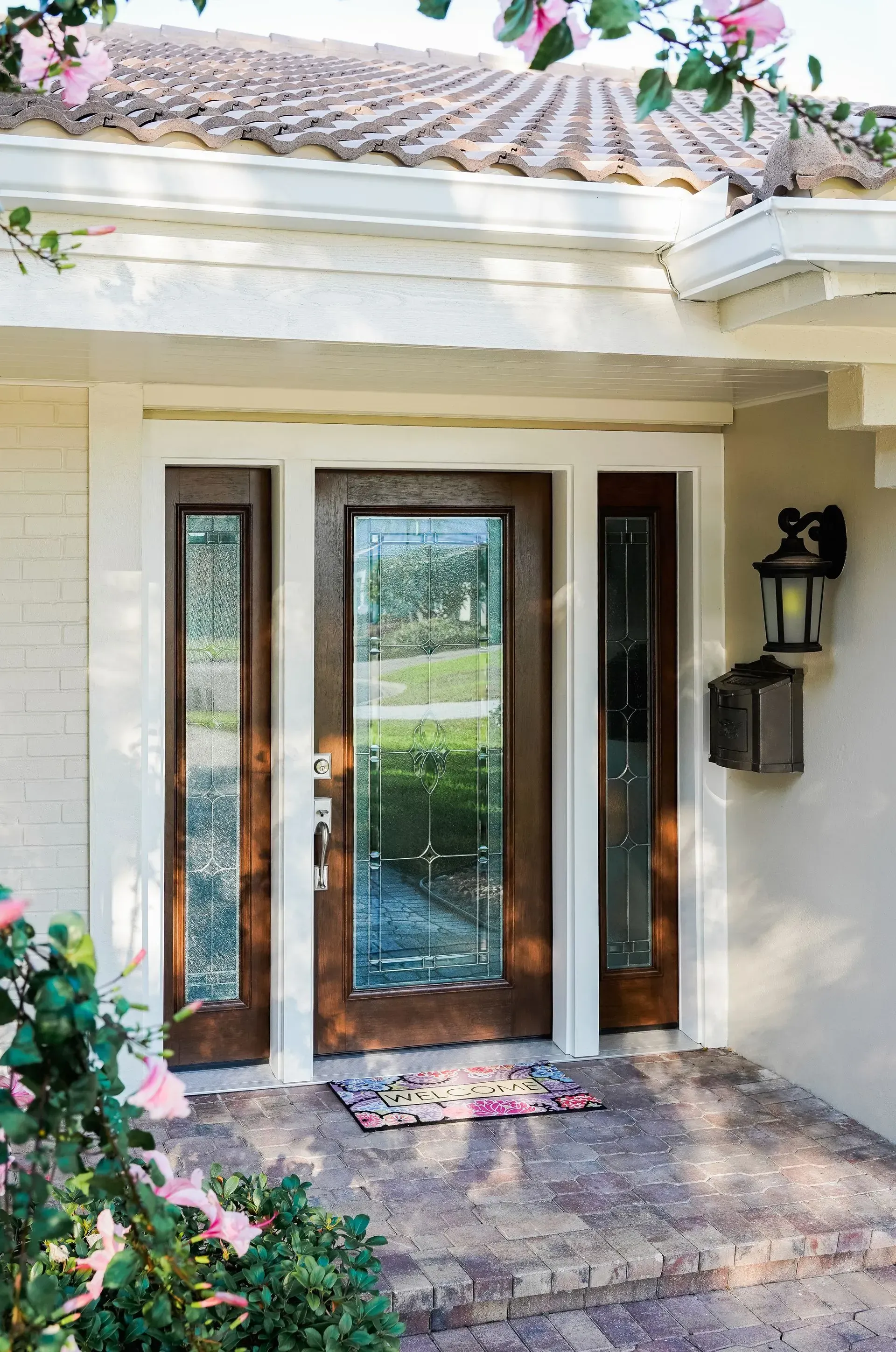 Front entryway with wood-framed glass door and sidelights, welcome mat, brick walkway, and a mailbox.