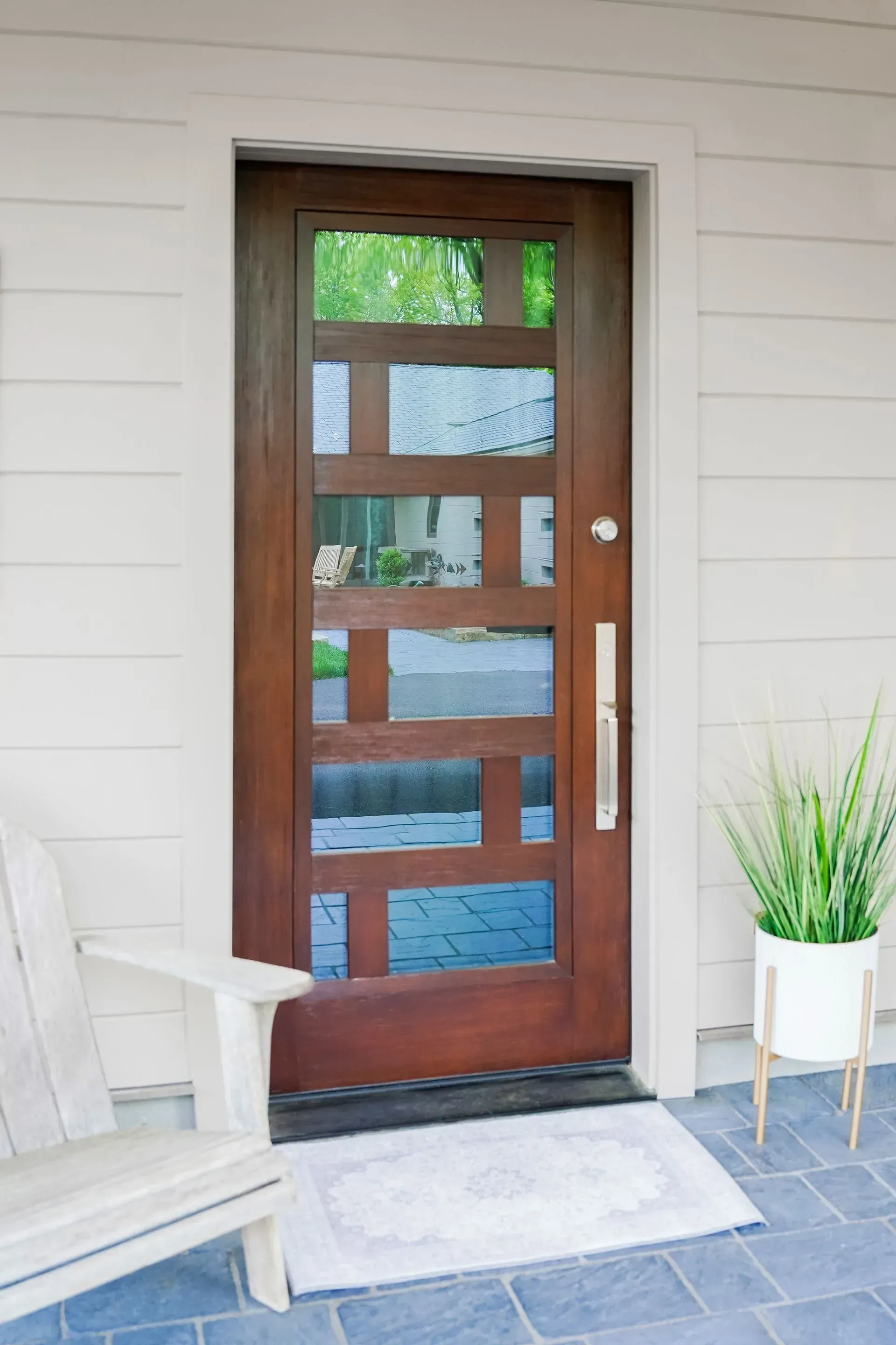 Brown wooden front door with geometric glass panels. Gray welcome mat, white chair, potted plant.