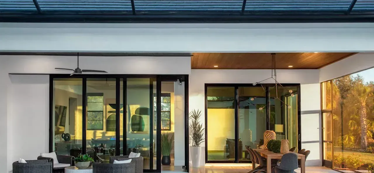 Outdoor patio with glass sliding doors, seating, and dining area. Sunlight streams through trees in the background.