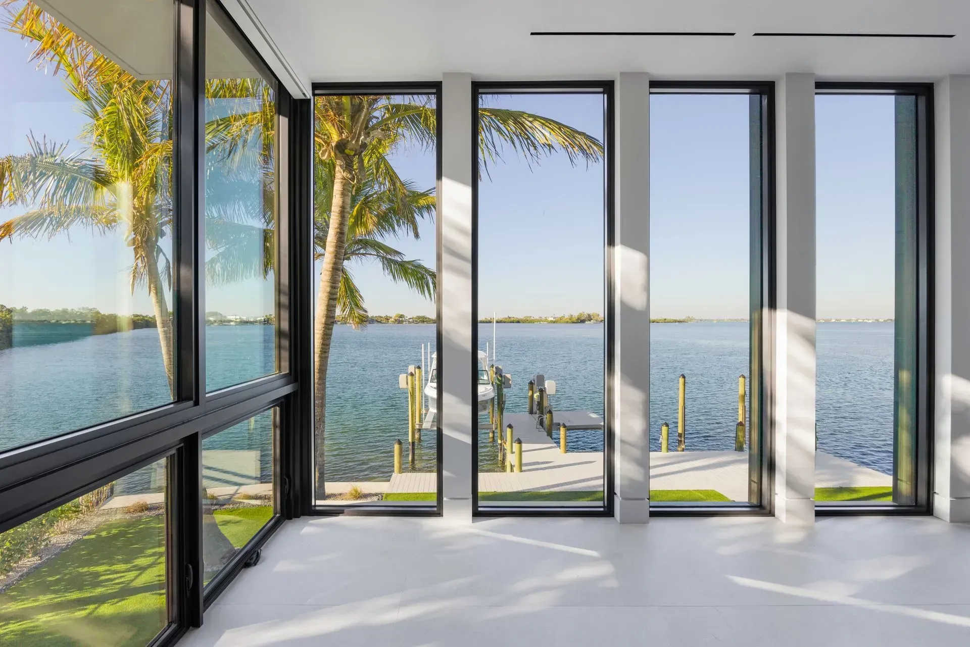 Interior room with large windows overlooking a waterfront view, including a dock, palm trees, and bright blue water.