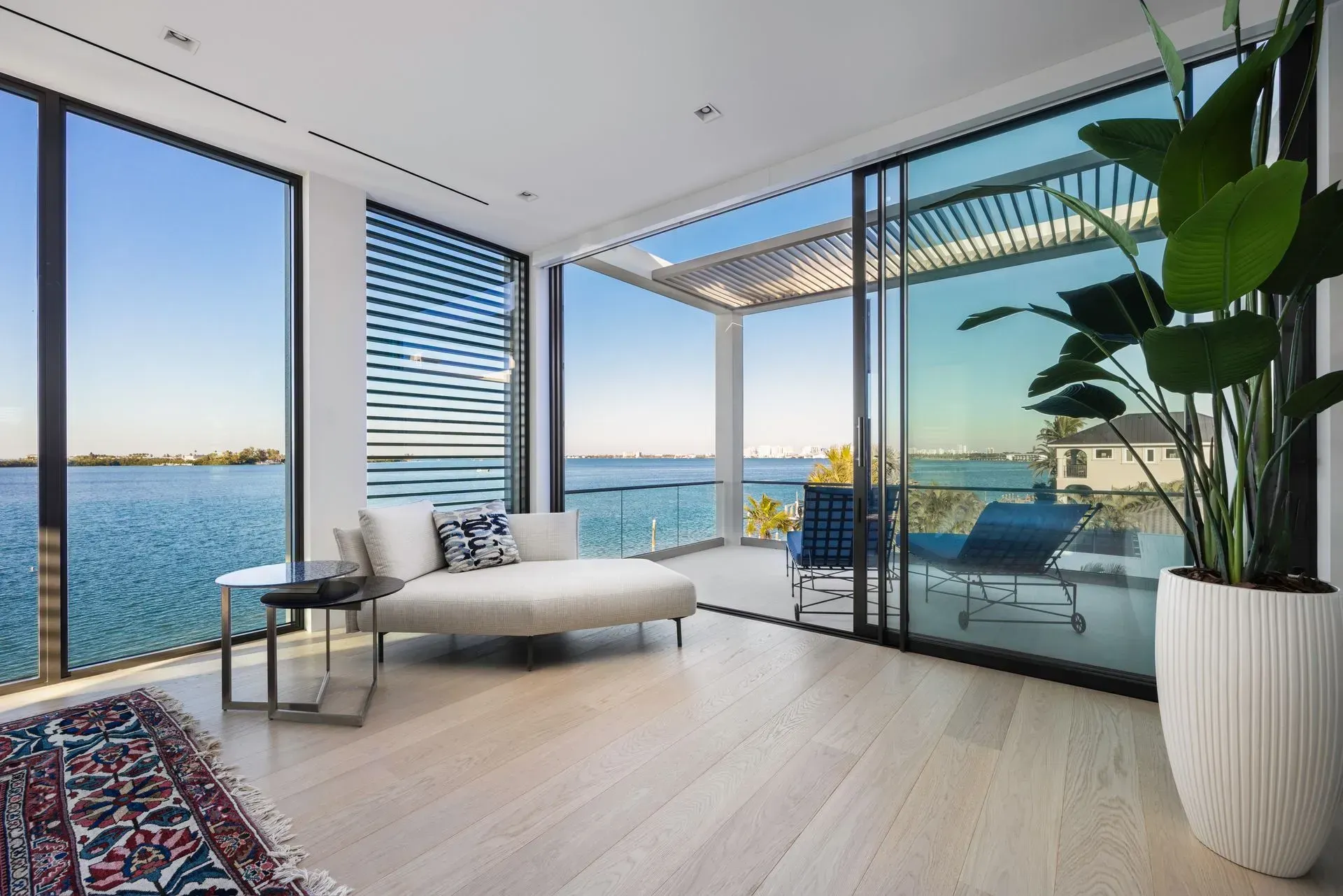 Bright room with large windows overlooking a blue body of water, with a modern white couch and a potted plant.
