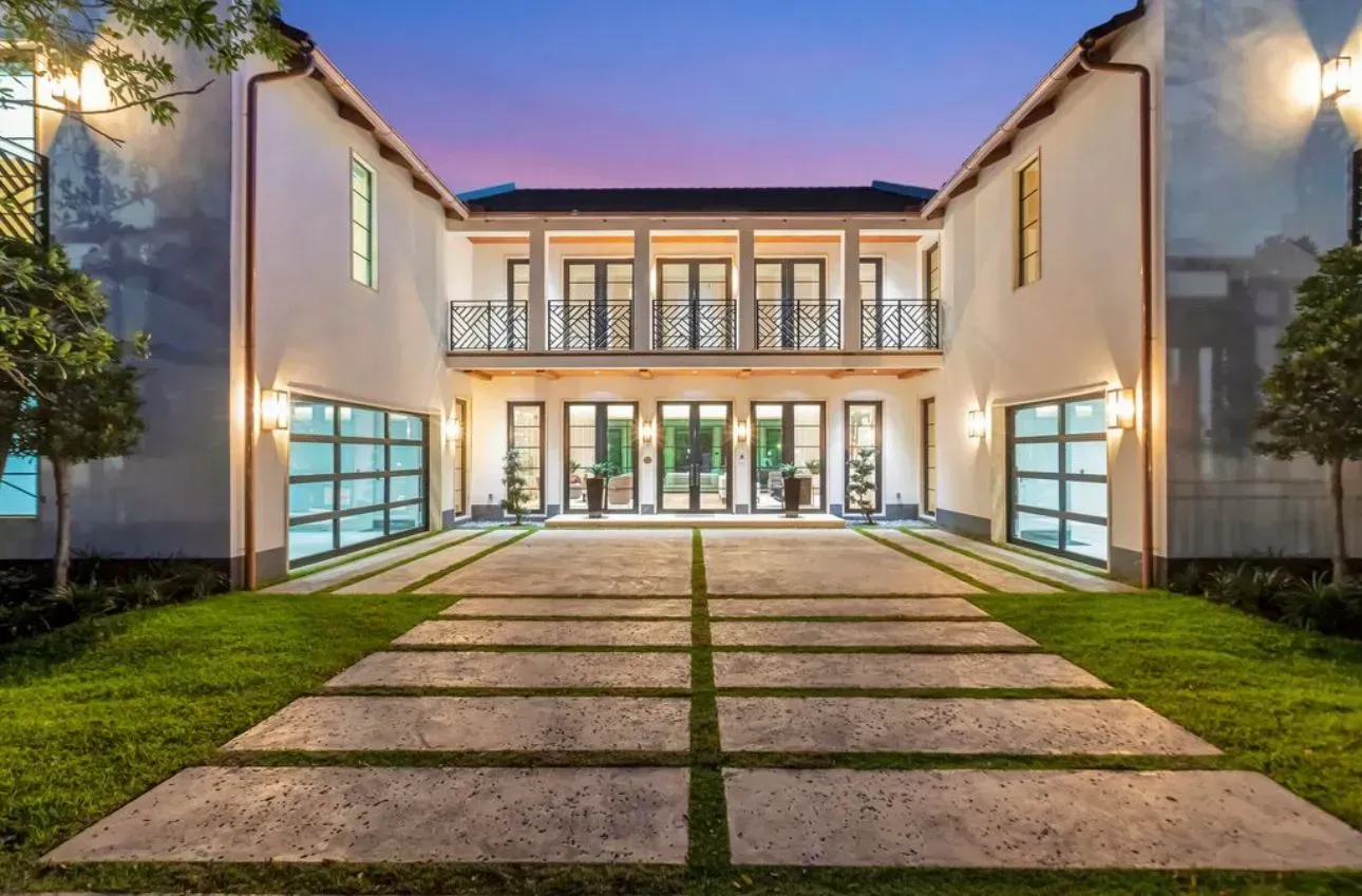 Two-story white house with glass garage doors, a stone driveway, and a balcony at twilight.