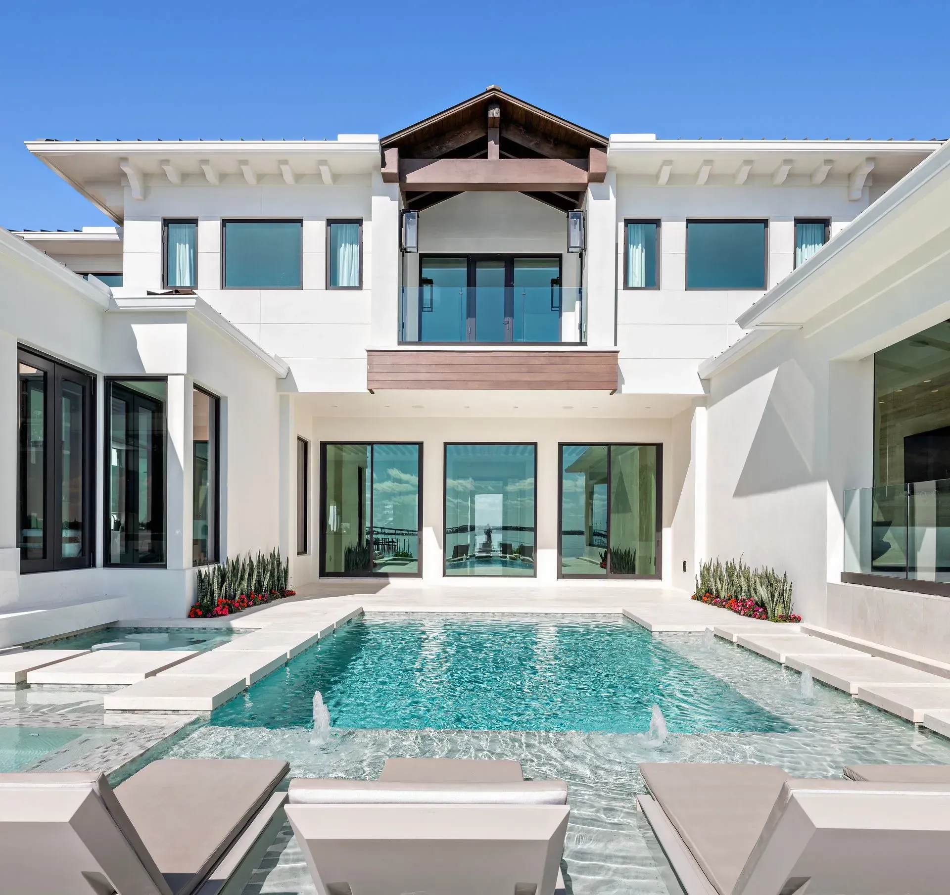 Luxury home exterior with pool and lounge chairs. White walls, blue sky, and large windows.