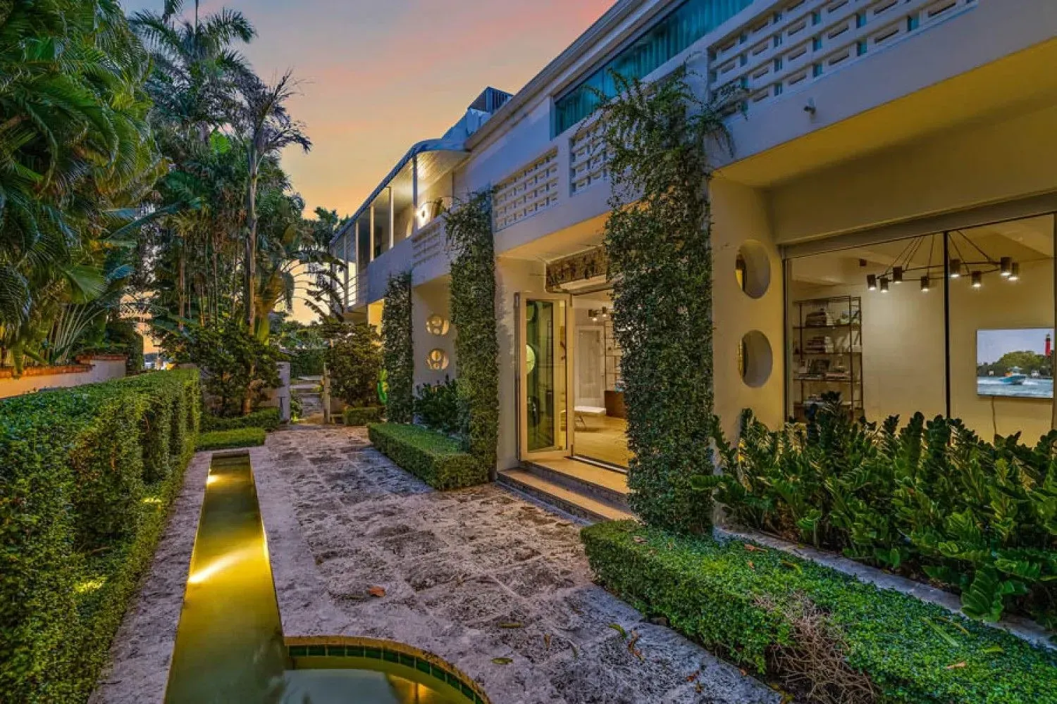 Exterior of a luxurious white building with a stone path, lush greenery, and a water feature at dusk.