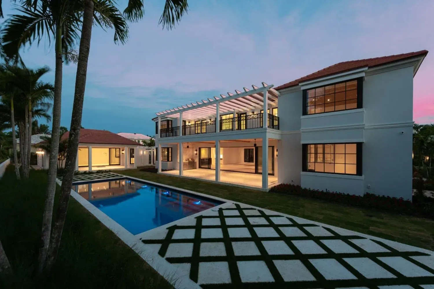 Modern house with pool at dusk, blue water, green grass, palm trees, and squared paving.
