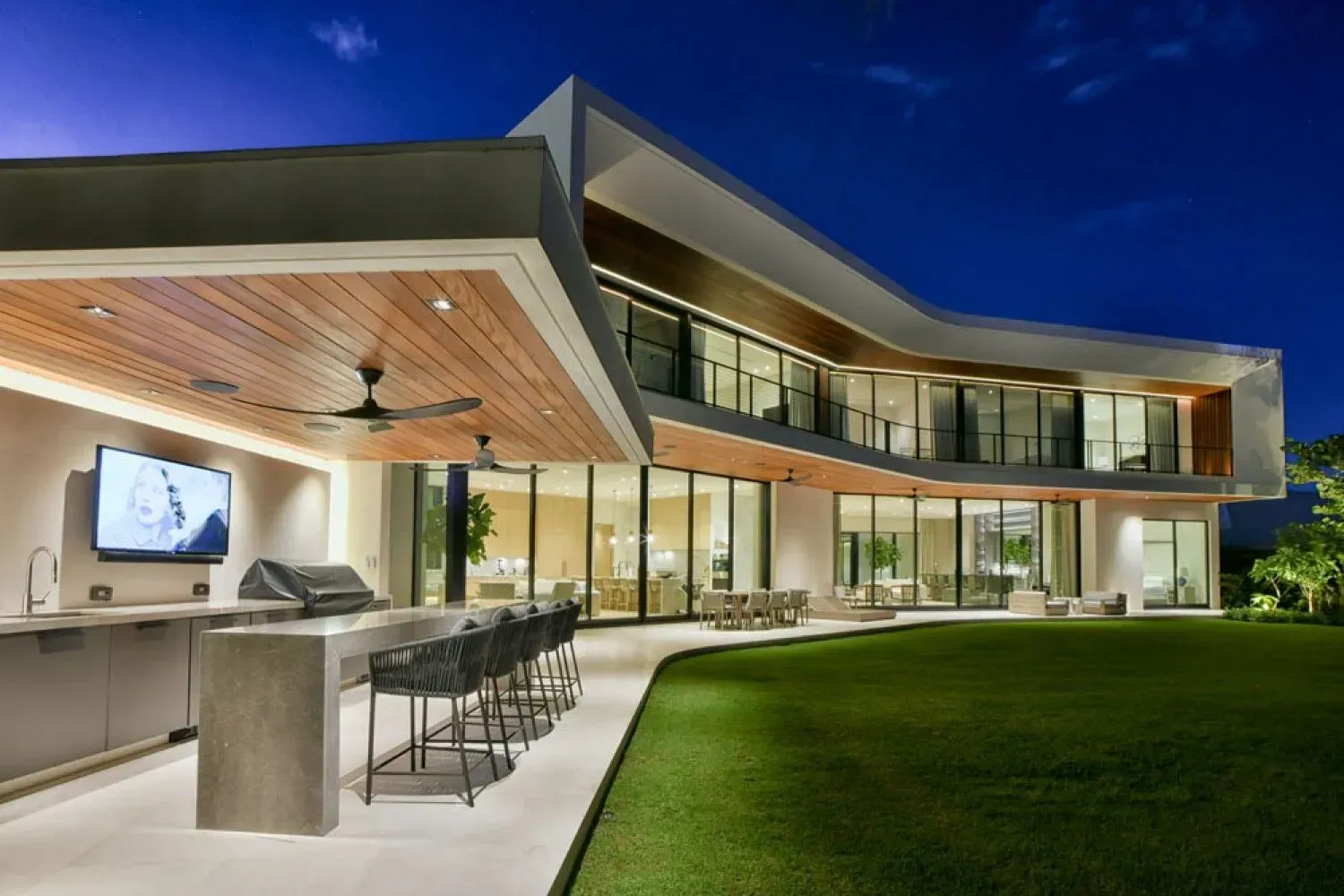 Modern house exterior with outdoor kitchen, lawn, and second-story balcony at dusk.