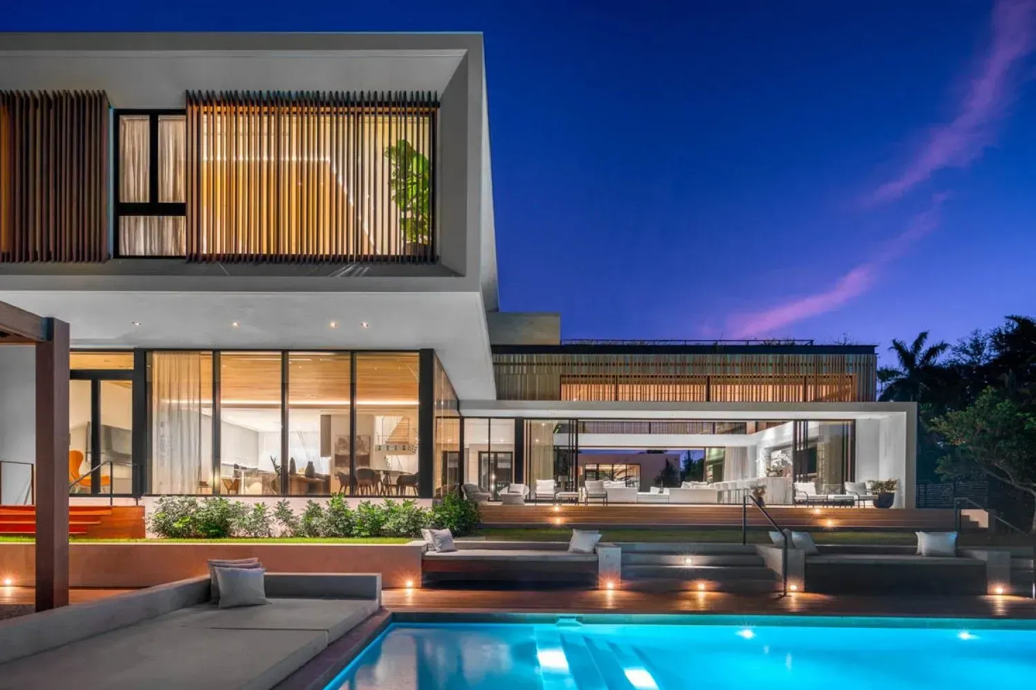 Modern home with pool, illuminated at dusk. Large windows, multiple levels, and outdoor seating.