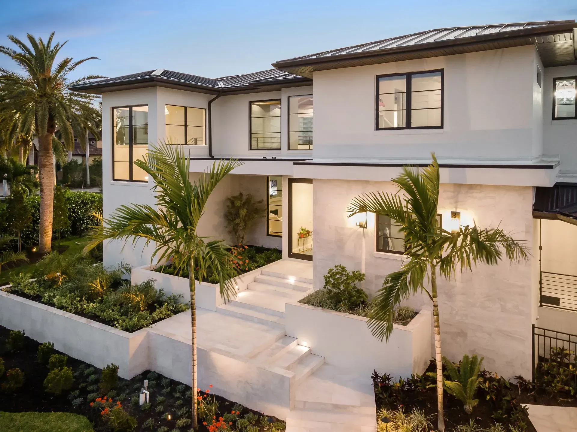 Modern white house with steps and palm trees. Black windows, black roof, and manicured landscaping.