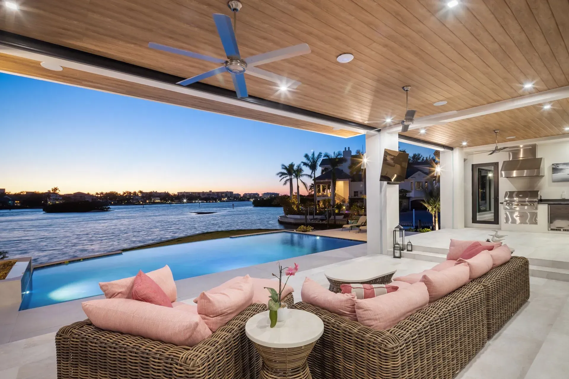 Patio with pool overlooking water at sunset. Wicker sofa with pink pillows, wooden ceiling, outdoor kitchen.