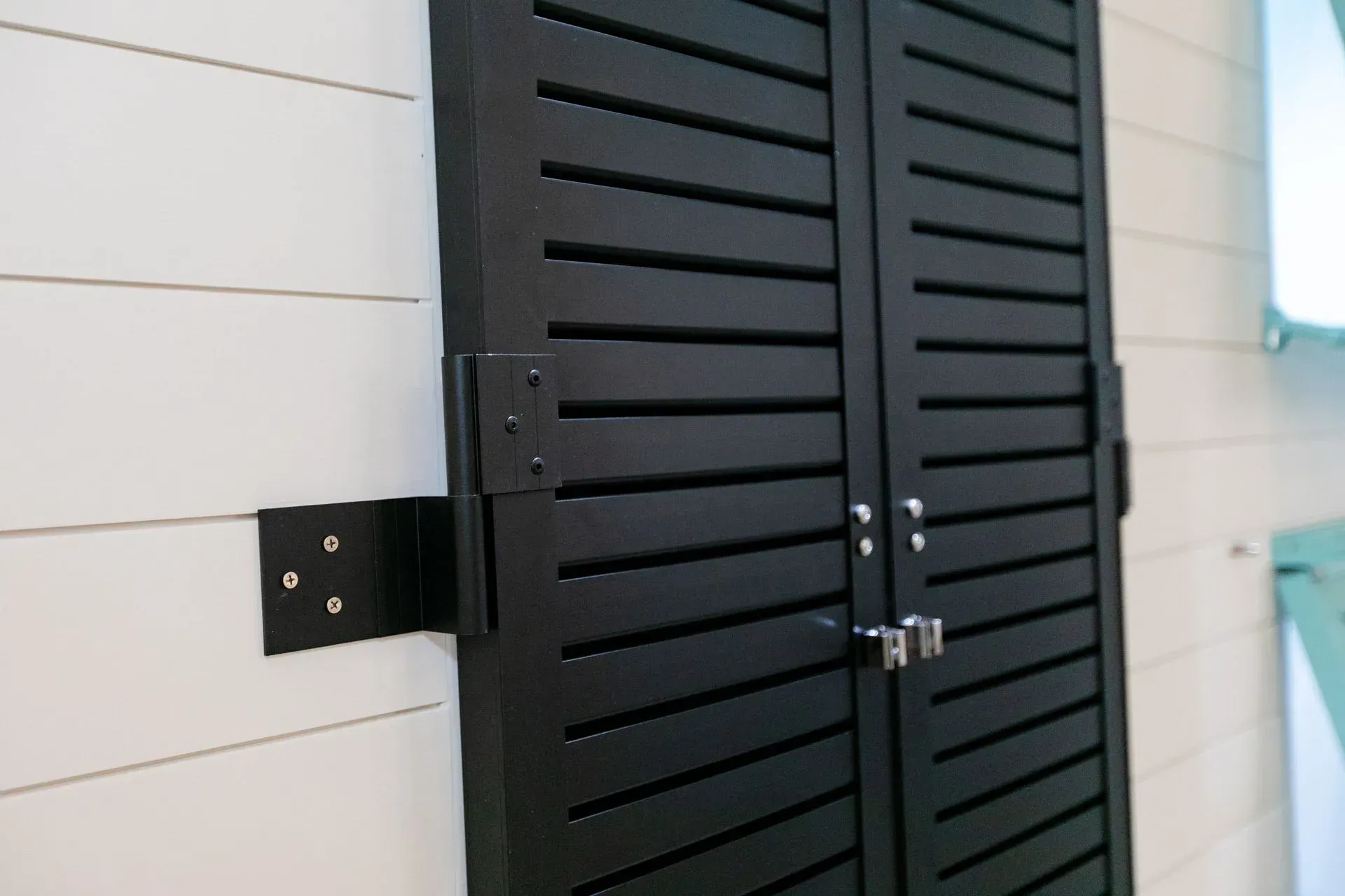 Black louvered shutters on a white shiplap wall. Metal hardware secures the shutters.
