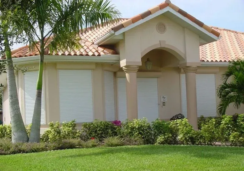 Beige house with white storm shutters, red tile roof, and green lawn.