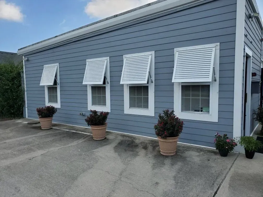 Blue building with white-framed windows, open white shutters, and potted plants on a concrete surface.