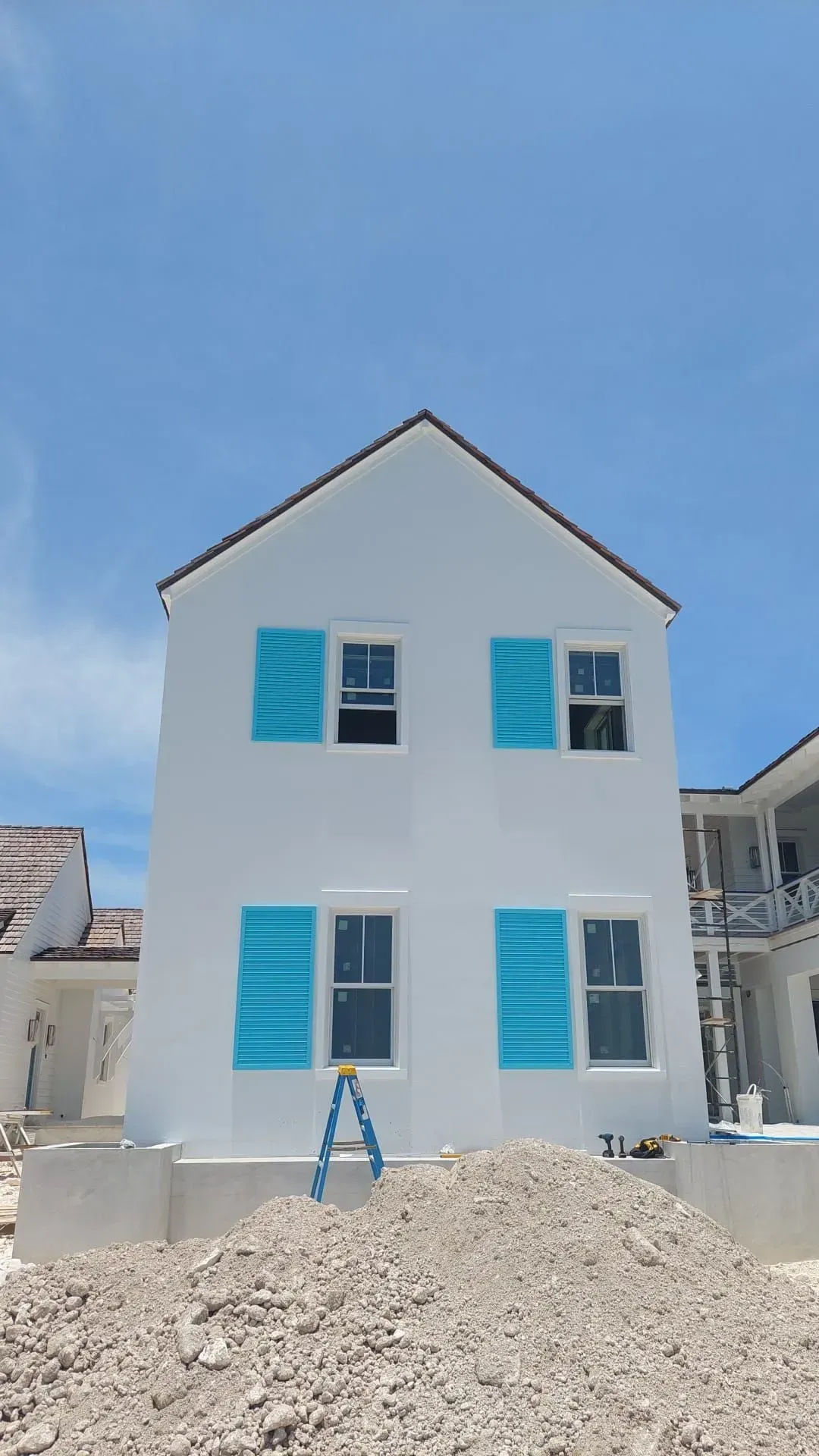 White two-story house with blue shutters, a brown roof, and a pile of dirt in front. Bright blue sky.