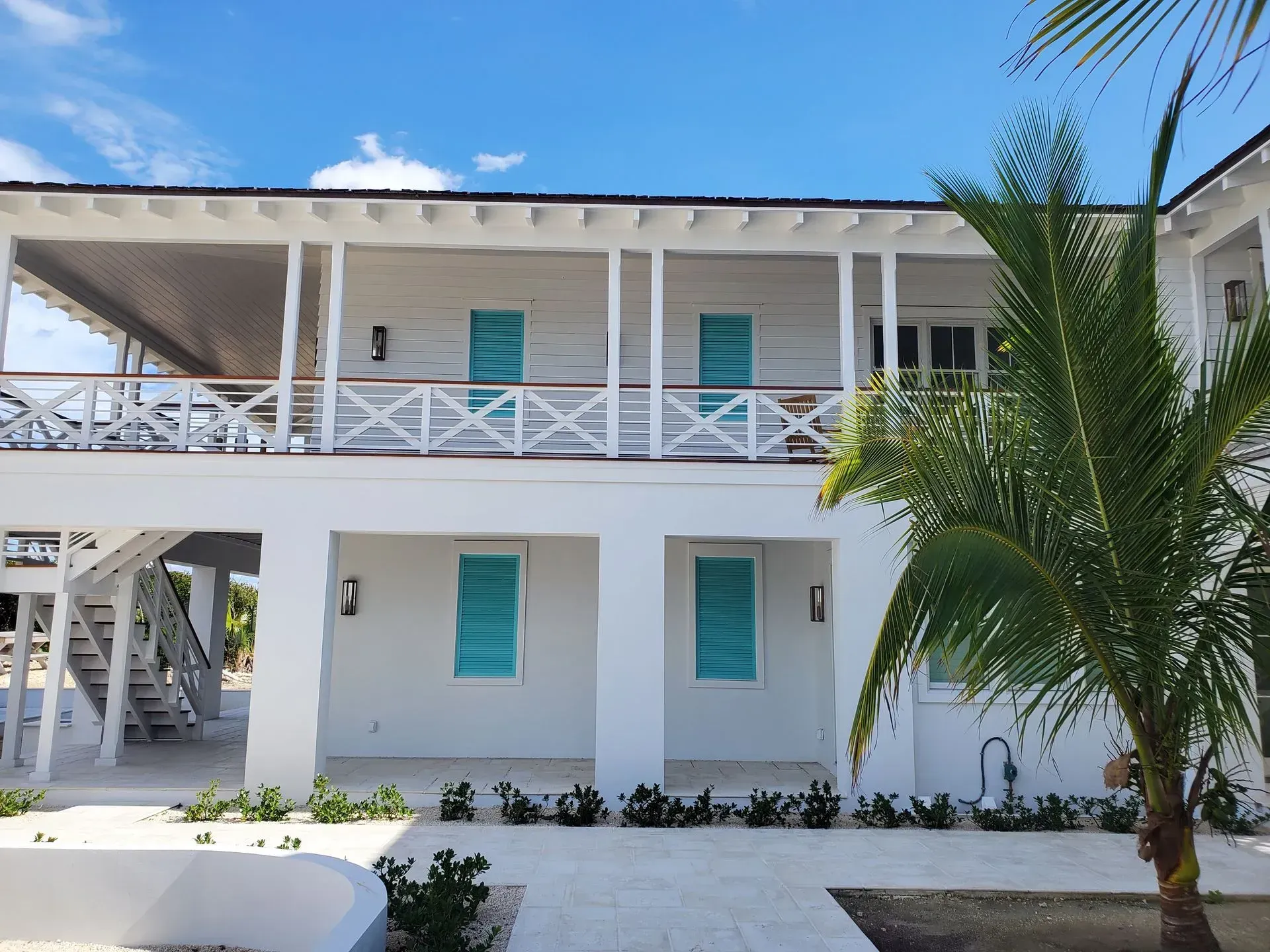 White two-story building with teal shutters, balconies, and palm trees against a blue sky.