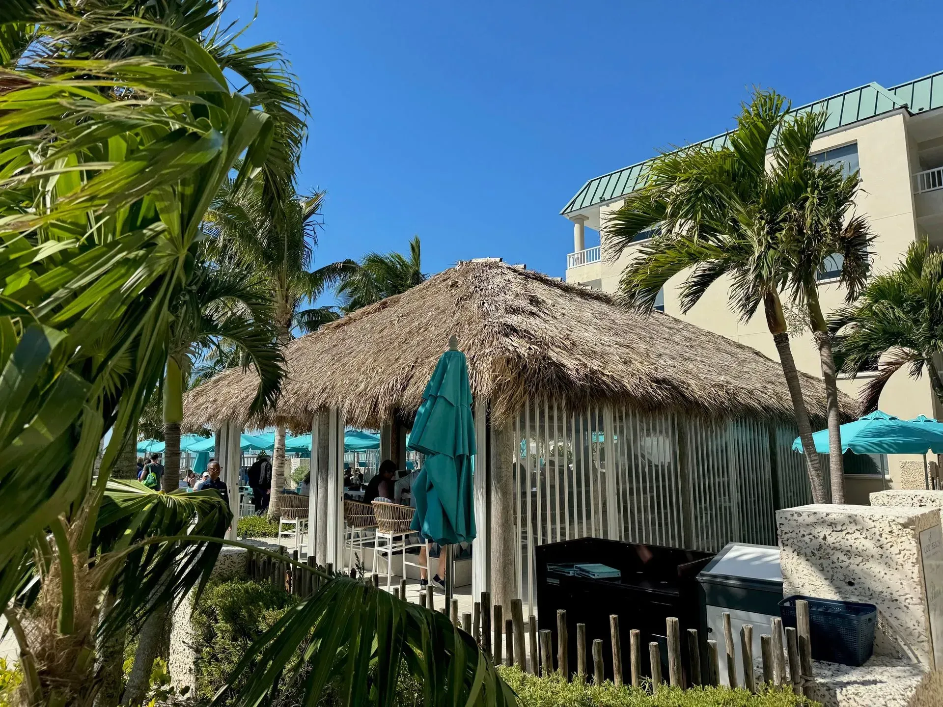 Thatched-roof cabana with teal umbrellas and palm trees under a bright blue sky.