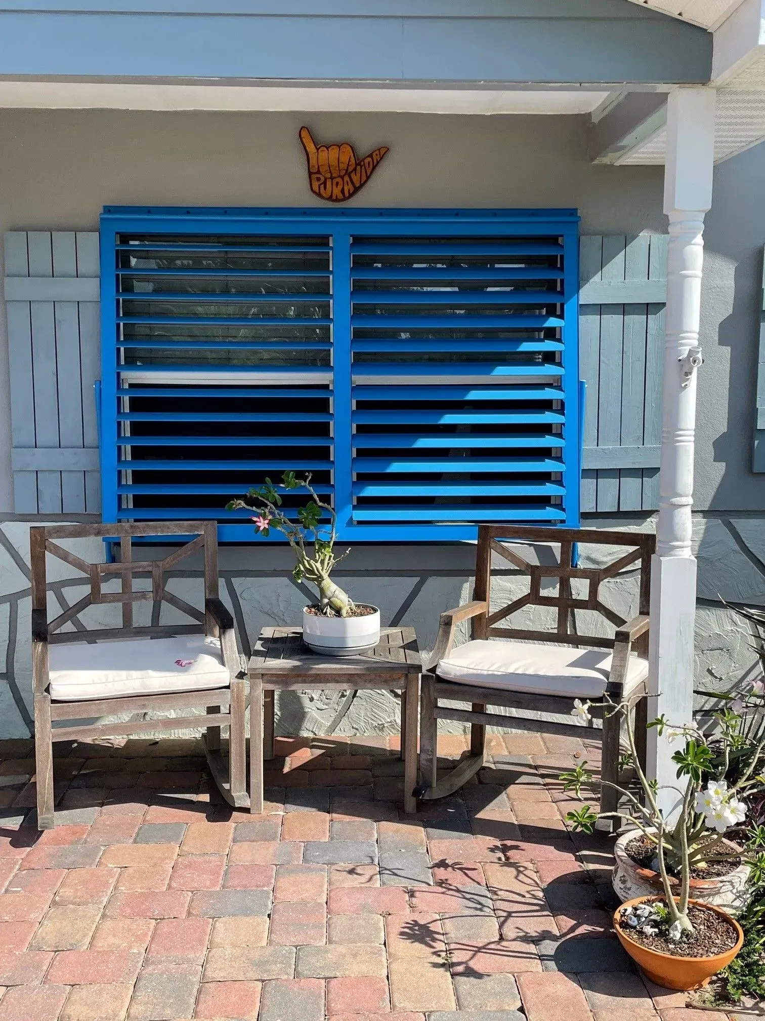 Two wooden chairs and a small table on a brick patio in front of a blue shuttered window.
