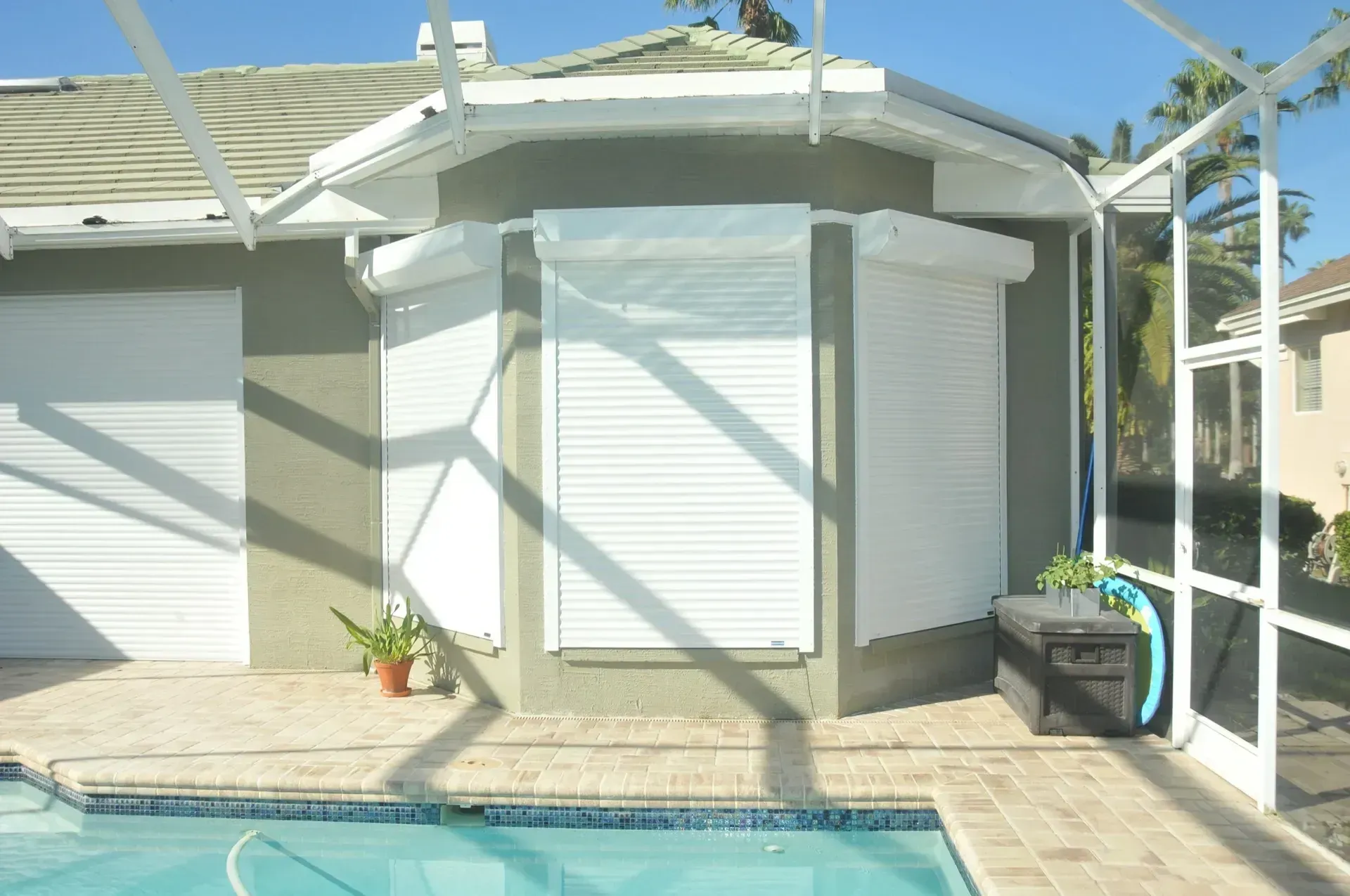 Exterior of a house with white hurricane shutters around a corner, adjacent to a pool with screen enclosure.